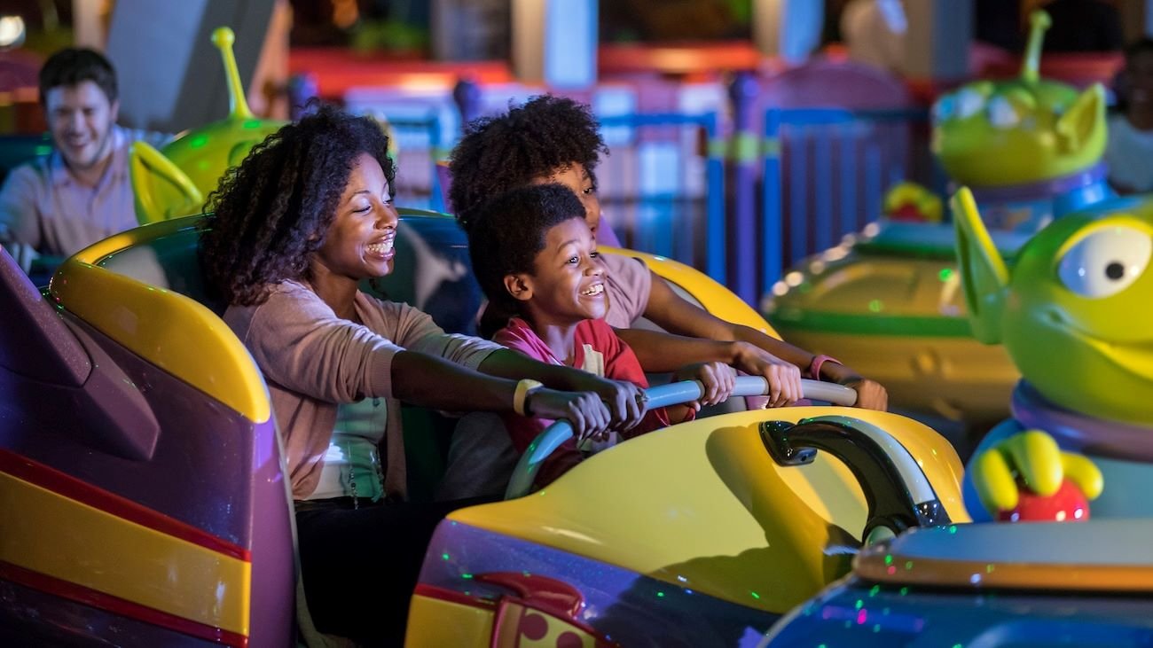 A woman and two children laughing as they ride a yellow and purple Alien Swirling Saucers vehicle at night, with green alien figures nearby