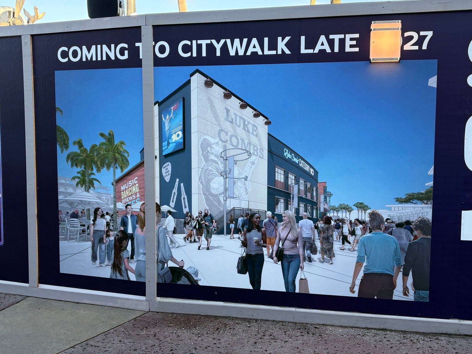 A construction wall panel reading Coming to CityWalk Late 2027 showing a rendering of a building with a white side wall painted with a large Luke Combs mural, crowds walking past in daylight