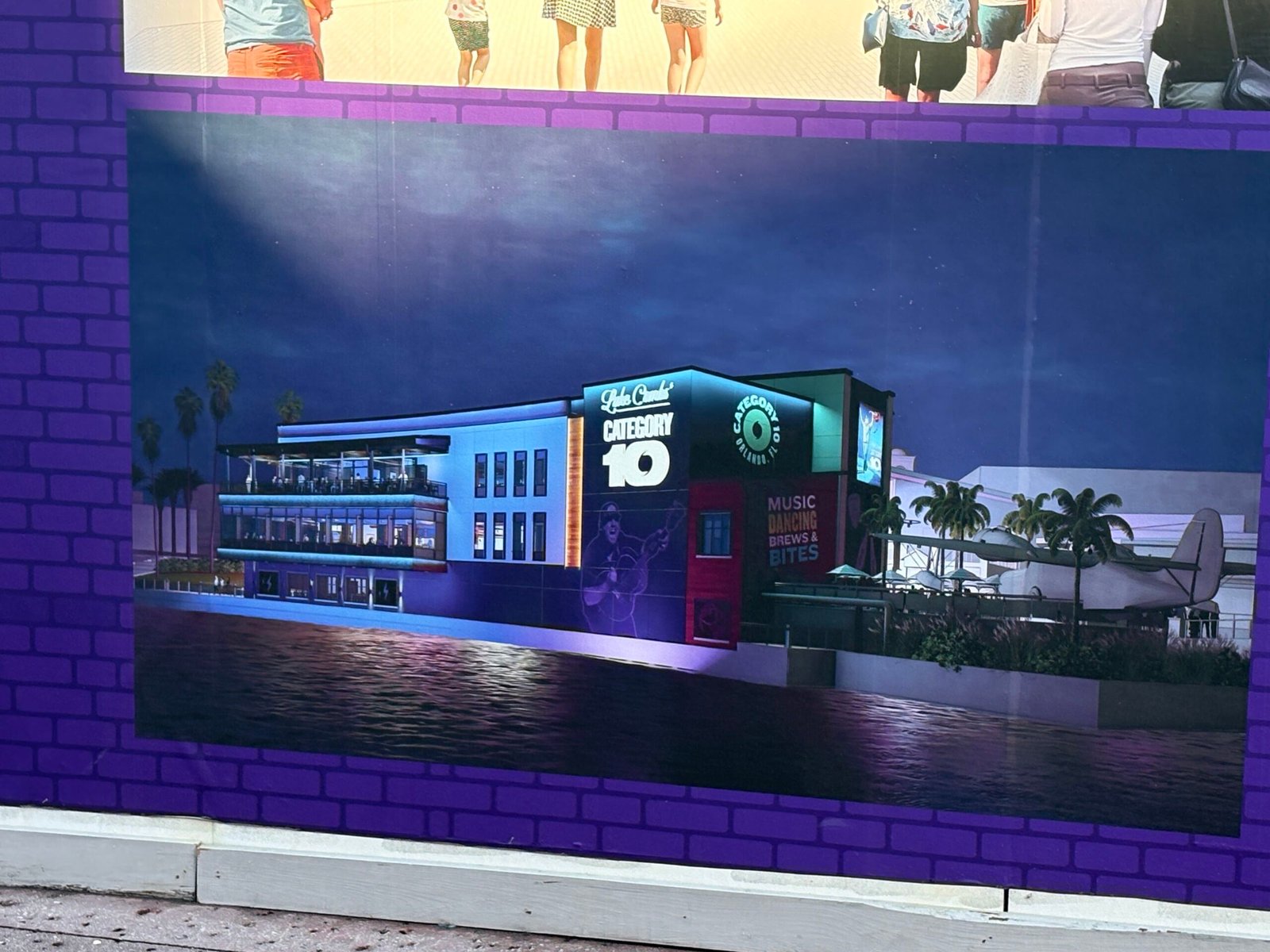 A construction wall panel showing a night rendering of the Category 10 building lit in blue and purple across the water, with a Music Dancing Brews and Bites sign and a seaplane to the right