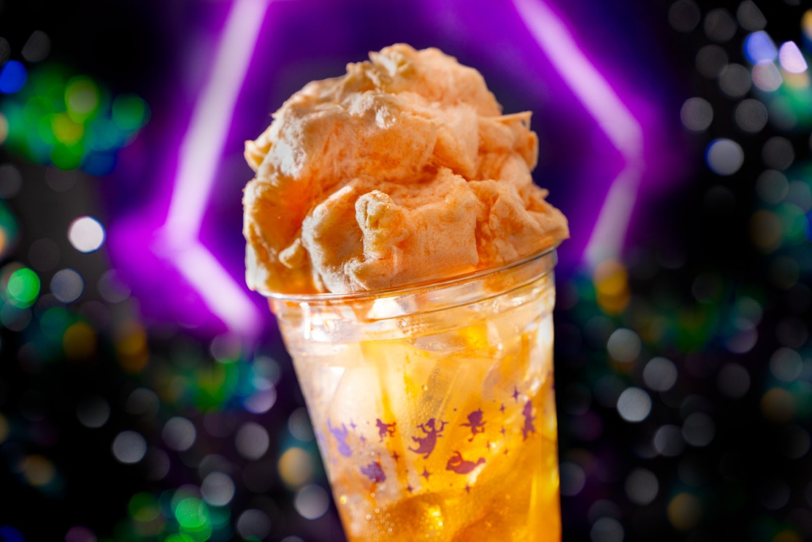 An orange drink in a clear cup topped with a large mound of orange cotton candy, against a purple neon light and colorful blurred lights