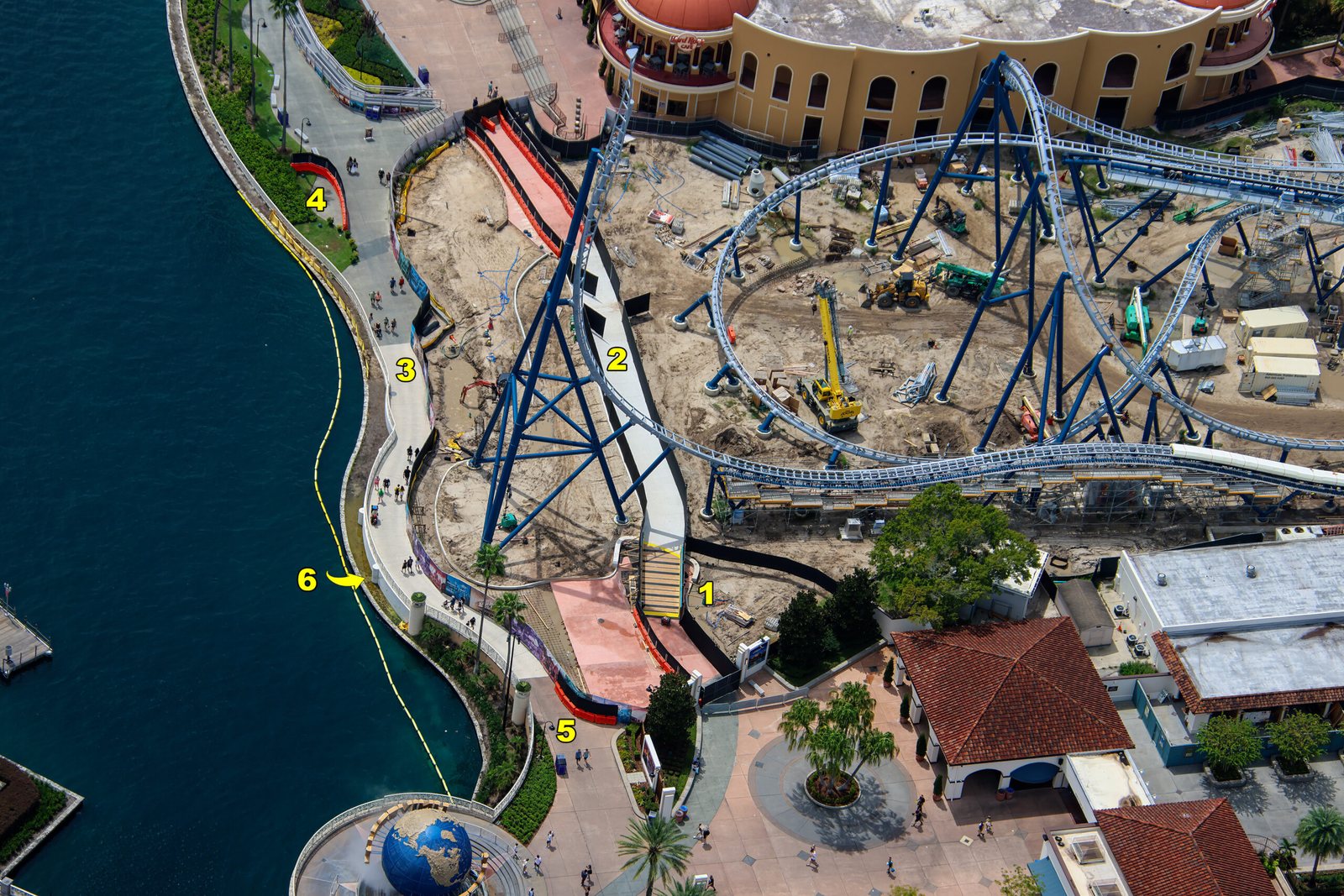 Aerial view of the lagoon edge with yellow numbers 1 through 6 marking a ramp, sidewalks and barricades beneath the tall blue spike supports of the coaster, with the Universal globe at the bottom