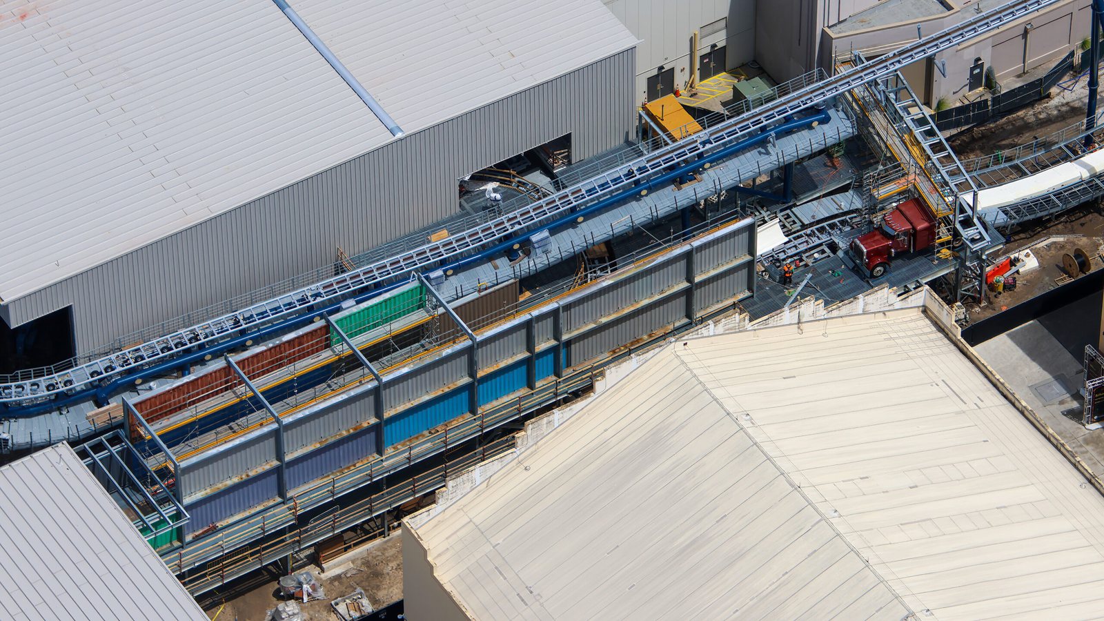 Aerial view of a long steel-framed wall clad in rust red, green, blue and gray corrugated panels beside elevated coaster track, with a burgundy semi truck cab on a platform at the far end