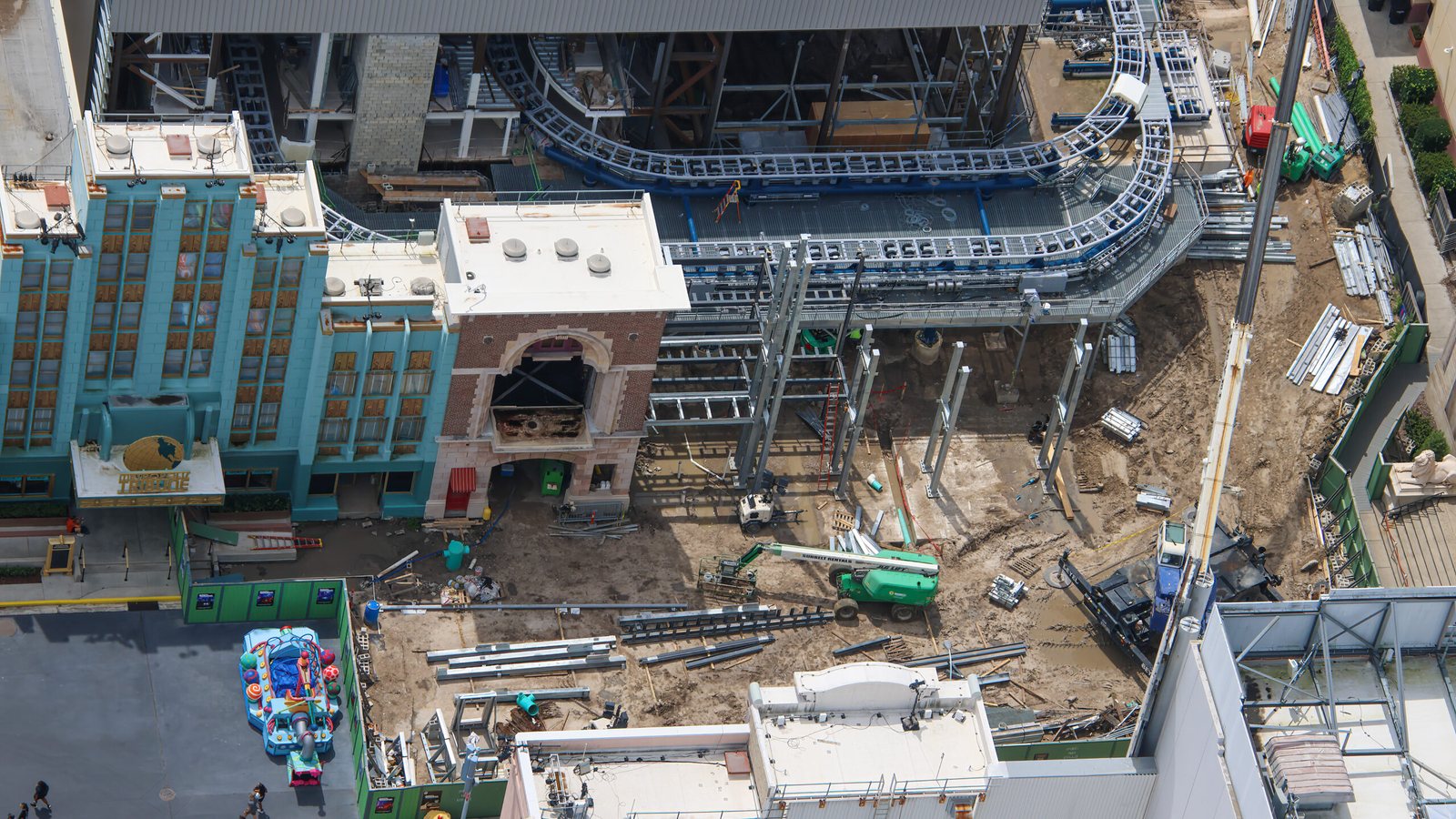Aerial view of the Ghostbusters firehouse facade with its brick arch beside a teal New York style building, with new steel columns rising on bare ground to the right and coaster track curving behind
