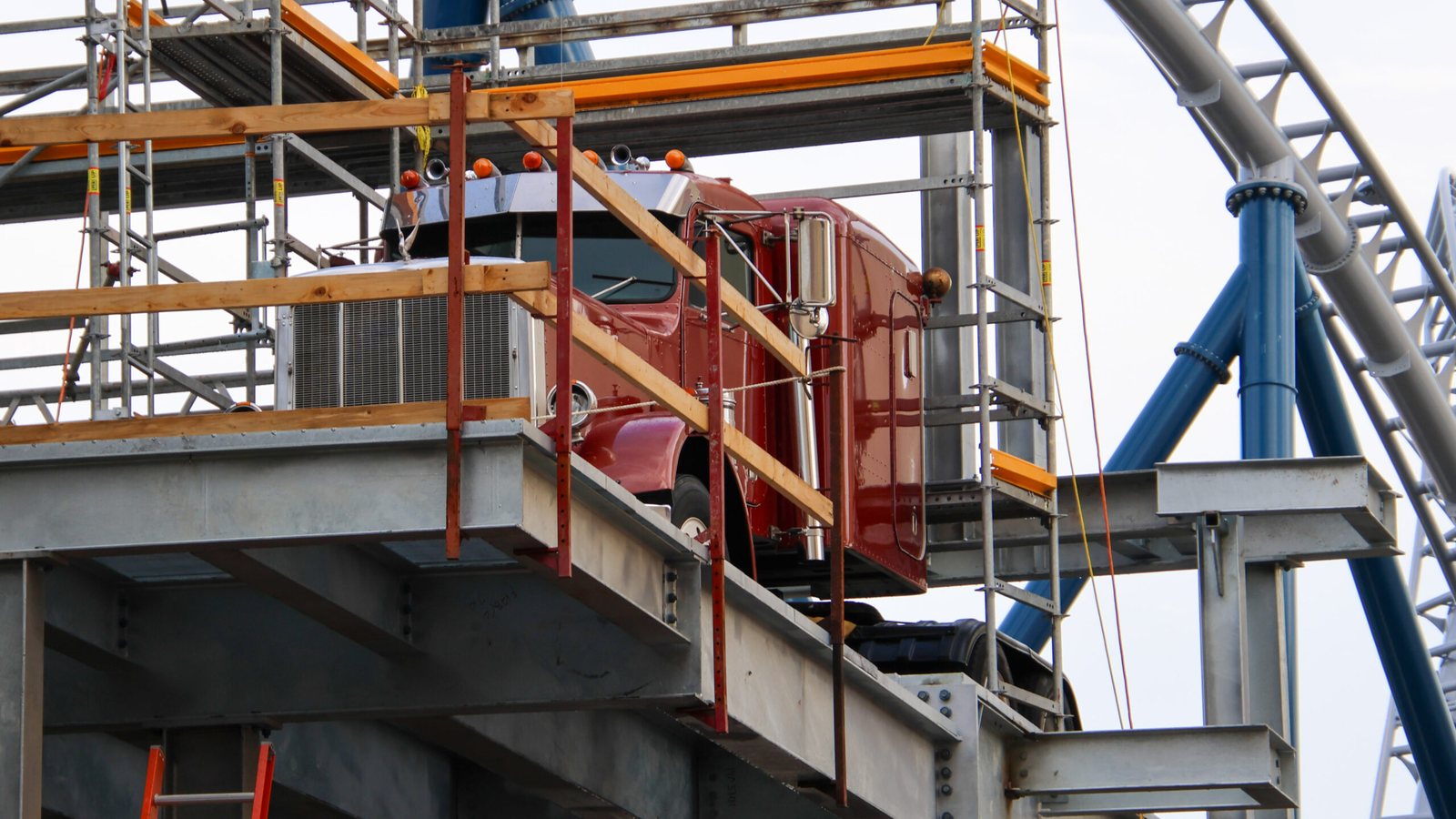 Close view of a burgundy semi truck cab with a chrome exhaust stack sitting on a steel platform surrounded by scaffolding, with blue coaster supports and track to the right