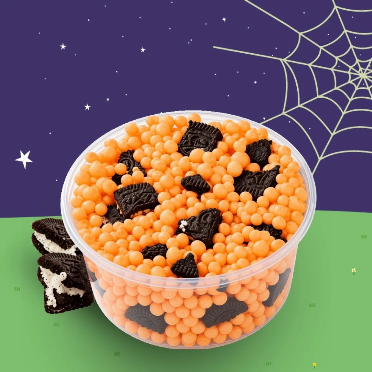 A clear cup filled with small orange ice cream beads and dark chocolate sandwich cookie pieces, against a starry sky and spider web