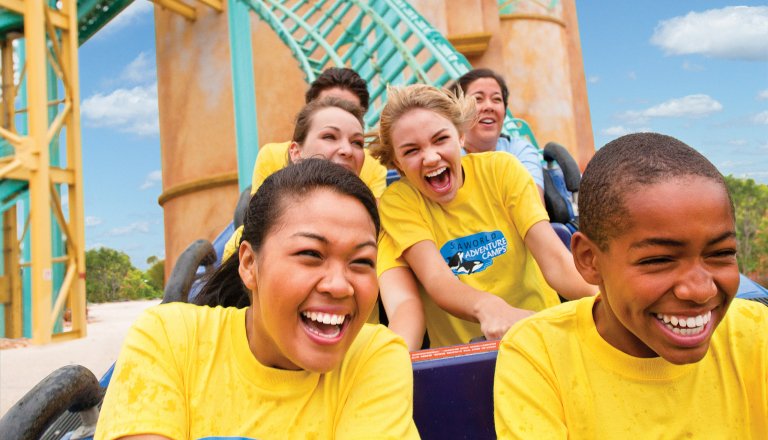 Laughing campers, most in yellow camp shirts and one reading SeaWorld Adventure Camps, ride a roller coaster with teal track and yellow supports behind them