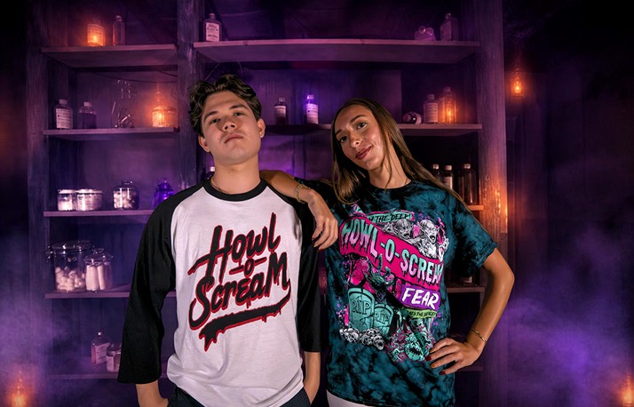 Two models in Howl-O-Scream shirts, one in a white and black raglan with the Howl-O-Scream script logo and one in a teal tie-dye shirt with the From the Deep design, in front of shelves of glowing bottles