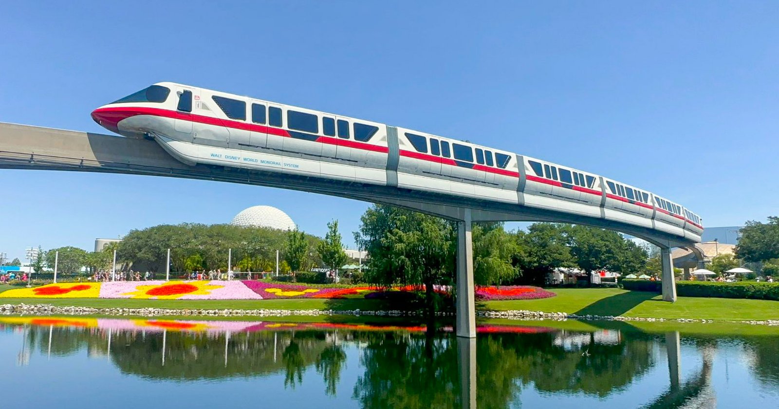 A white monorail with a red stripe curves over a pond and flower beds at EPCOT, with Spaceship Earth behind the trees