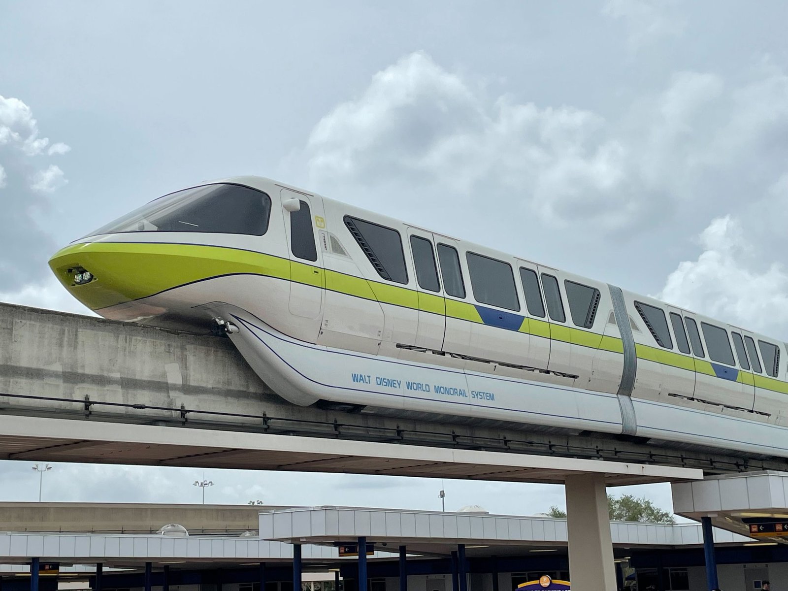 A white Walt Disney World monorail with a lime green stripe on an elevated concrete beam above covered walkways, under a cloudy sky