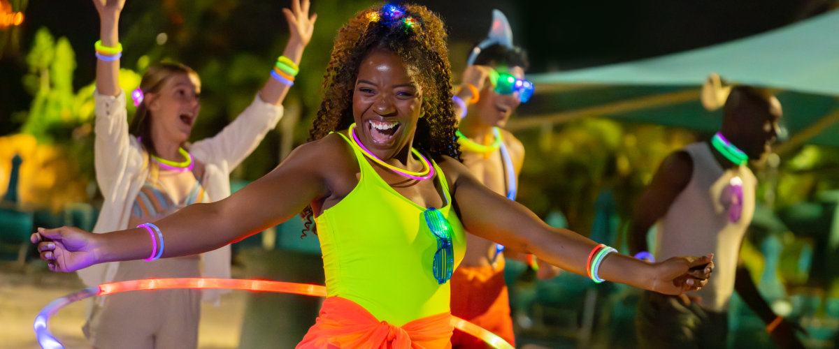 Guests at a night event at Aquatica Orlando wearing glow bracelets and necklaces, with a woman in a neon yellow swimsuit and orange wrap in the foreground spinning a light-up hula hoop around her waist
