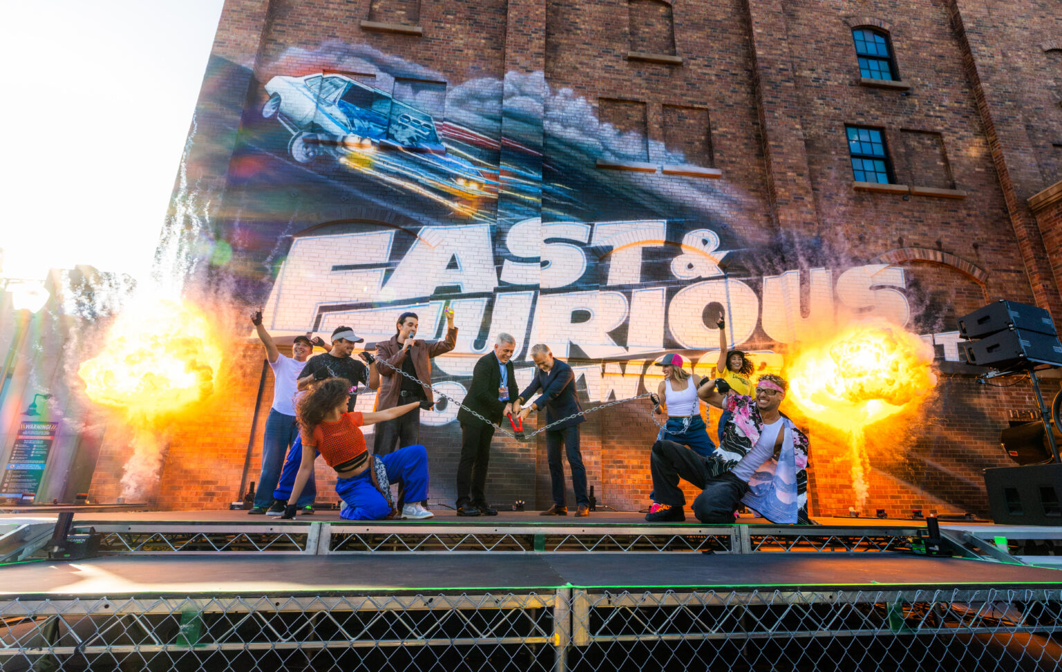 The Hollywood Drift opening ceremony: two men in suits cutting a chain with red bolt cutters on a stage, flanked by dancers with raised arms, two tall flame bursts on either side, in front of a brick building painted with a large Fast and Furious Hollywood Drift mural of a car mid-air