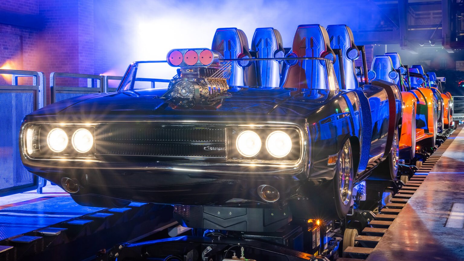 A Fast and Furious Hollywood Drift ride train on track in a station lit blue, led by a black Dodge Charger with an exposed chrome supercharger on the hood and four round headlights, with an orange car visible behind it and brick walls on either side