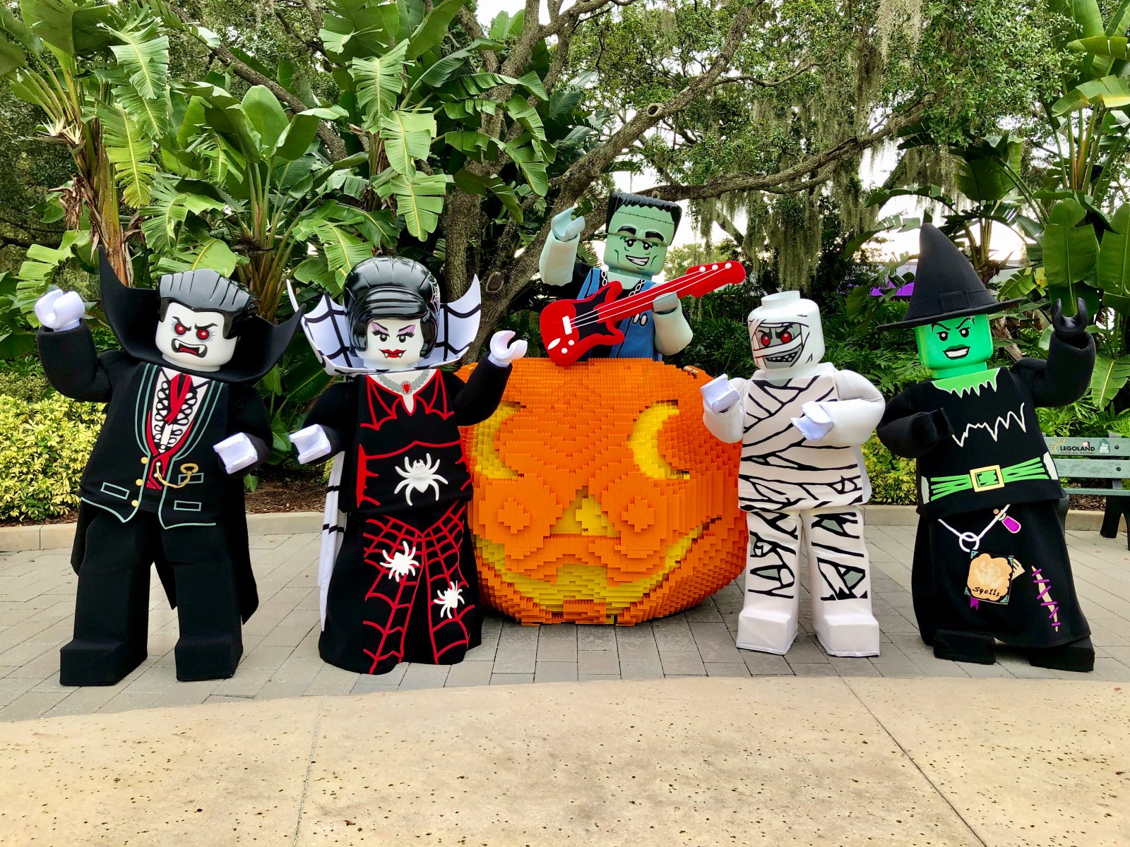 Five costumed LEGO Monster characters around a giant orange LEGO brick jack-o-lantern: a vampire, a woman in a black spiderweb gown, a green monster standing behind the pumpkin with a red guitar resting on top of it, a mummy and a green-faced witch in a pointed hat
