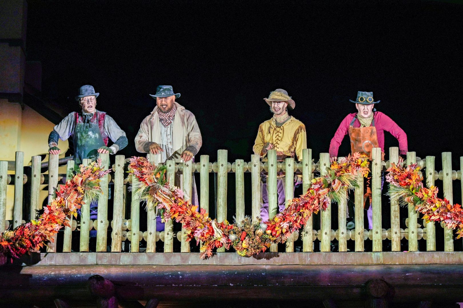 Four performers in weathered frontier costumes and ghostly pale makeup, wearing hats, two of them in work aprons, standing behind a wooden fence railing strung with autumn leaf garland on a dark stage at night