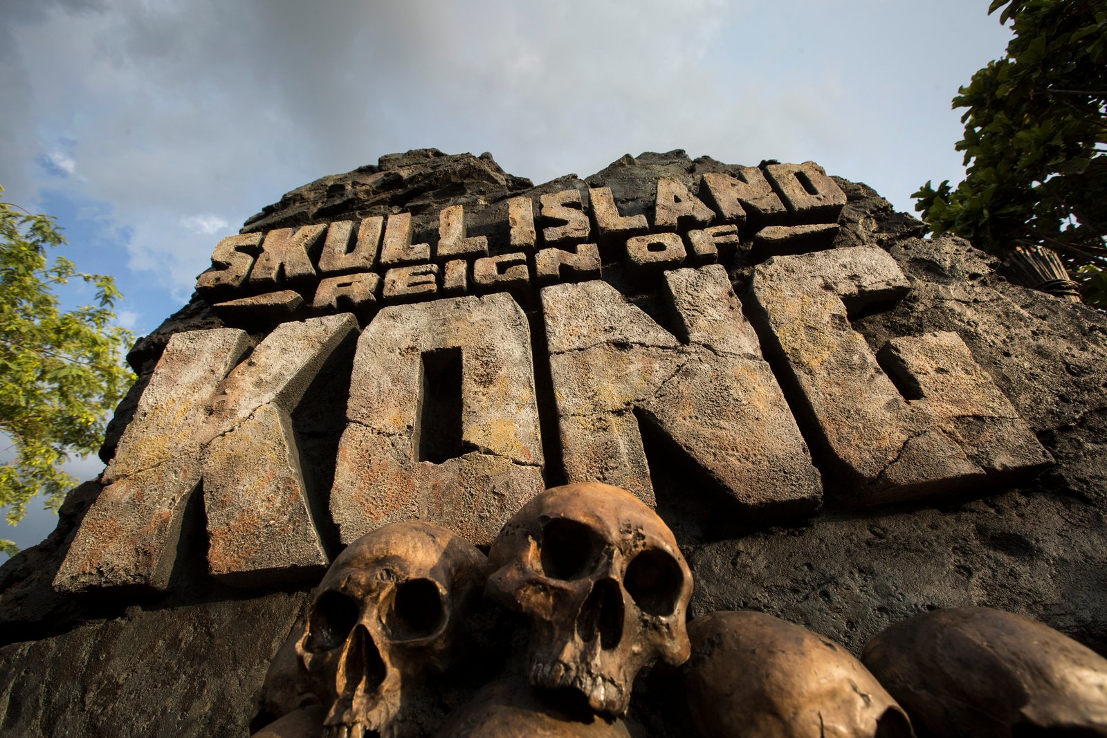 The carved stone Skull Island: Reign of Kong marquee with bronze skulls piled in front of it, against a partly cloudy sky