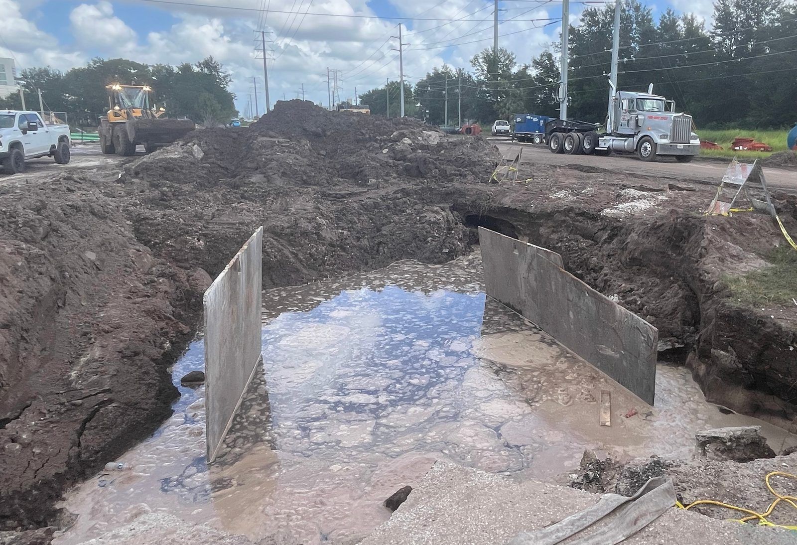 A large excavated pit filled with murky, foamy water between two steel trench plates, with piles of dirt, a front loader, a white pickup and a semi truck in the background