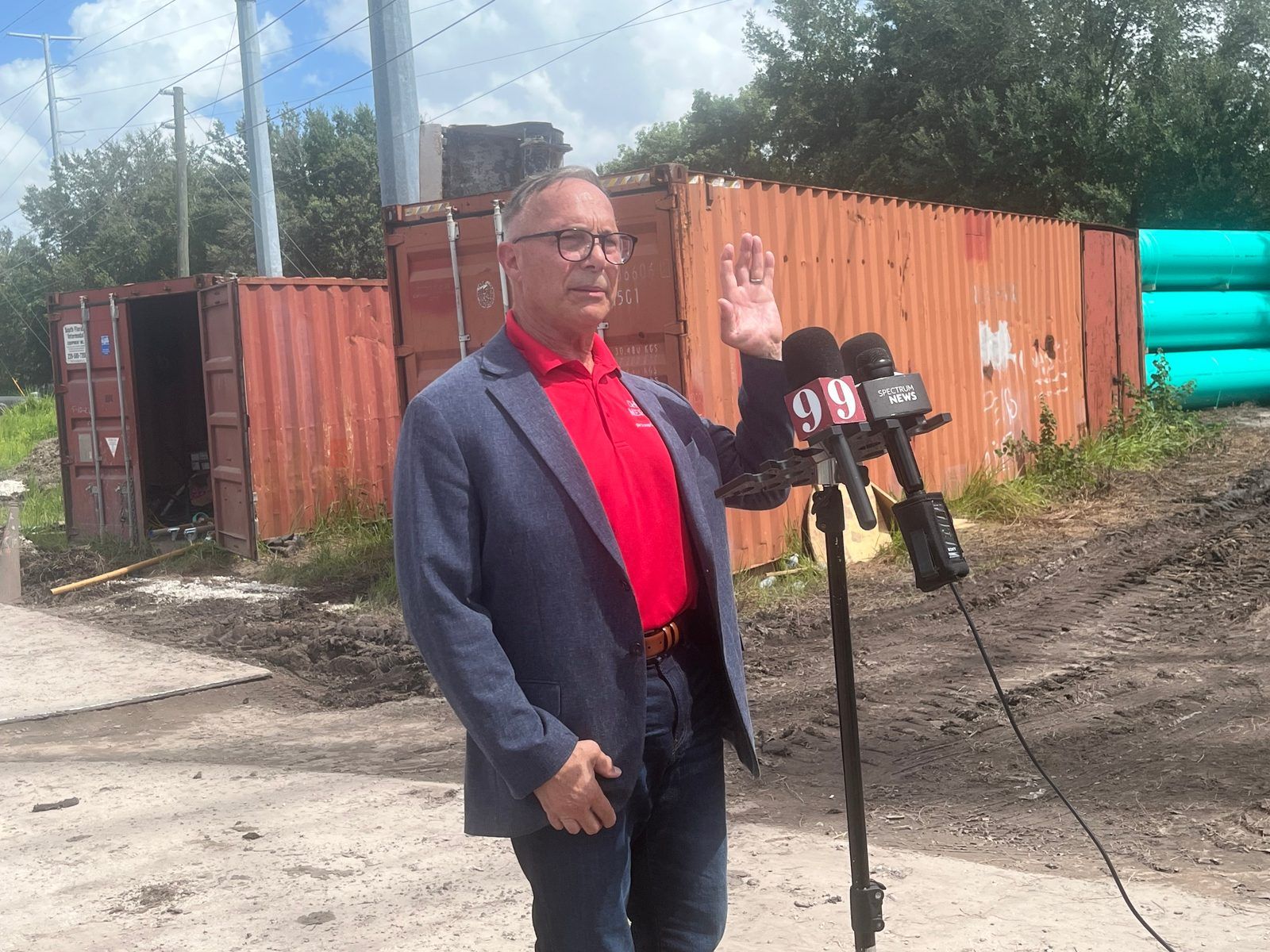 A man with glasses in a blue blazer and red polo shirt speaks into microphones from Channel 9 and Spectrum News, raising one hand, in front of rusted orange shipping containers