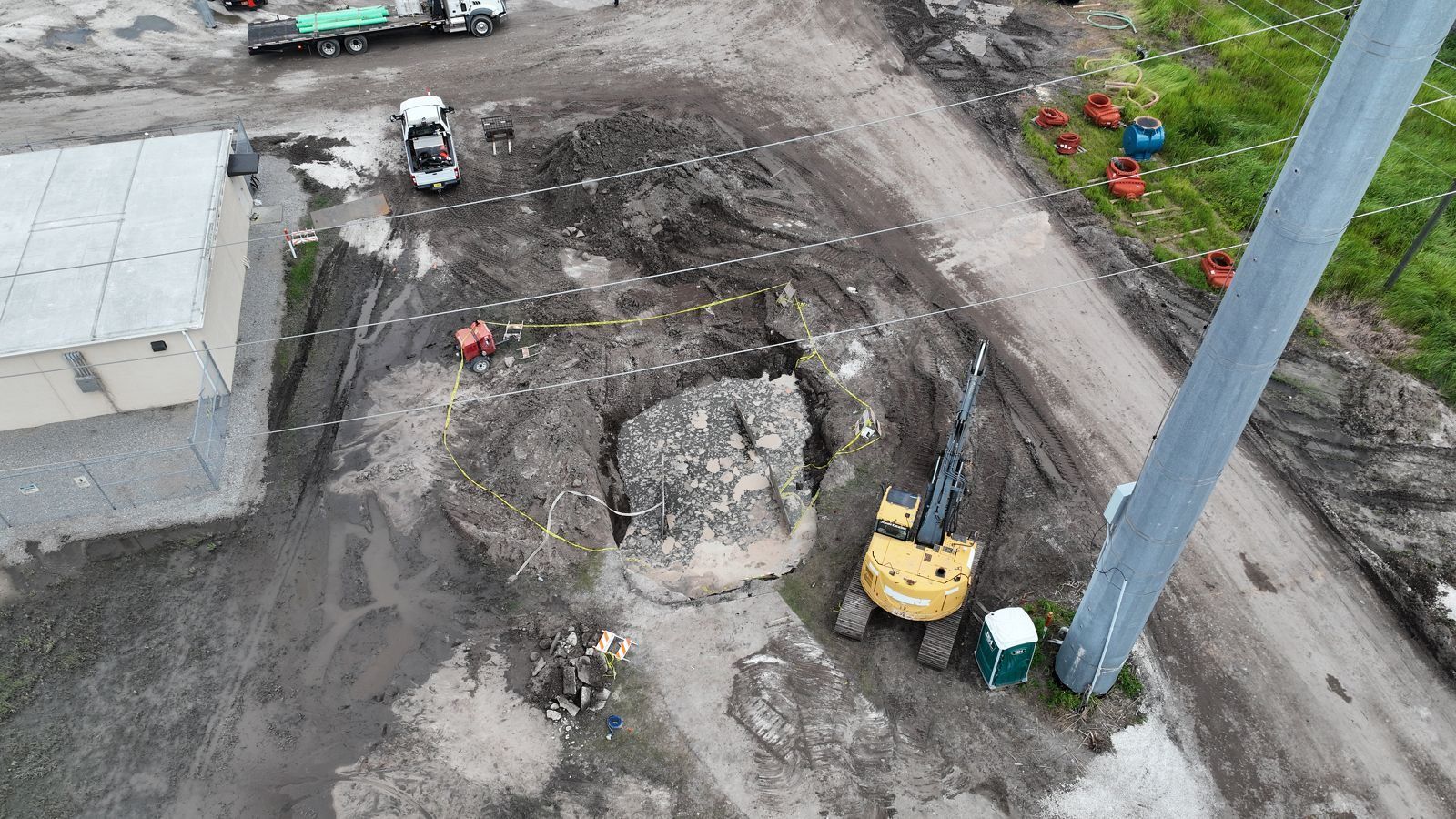 Aerial view of a muddy excavation beside a small beige utility building, with a yellow excavator, a portable toilet, a pickup truck and a flatbed trailer around a large hole ringed with yellow caution tape