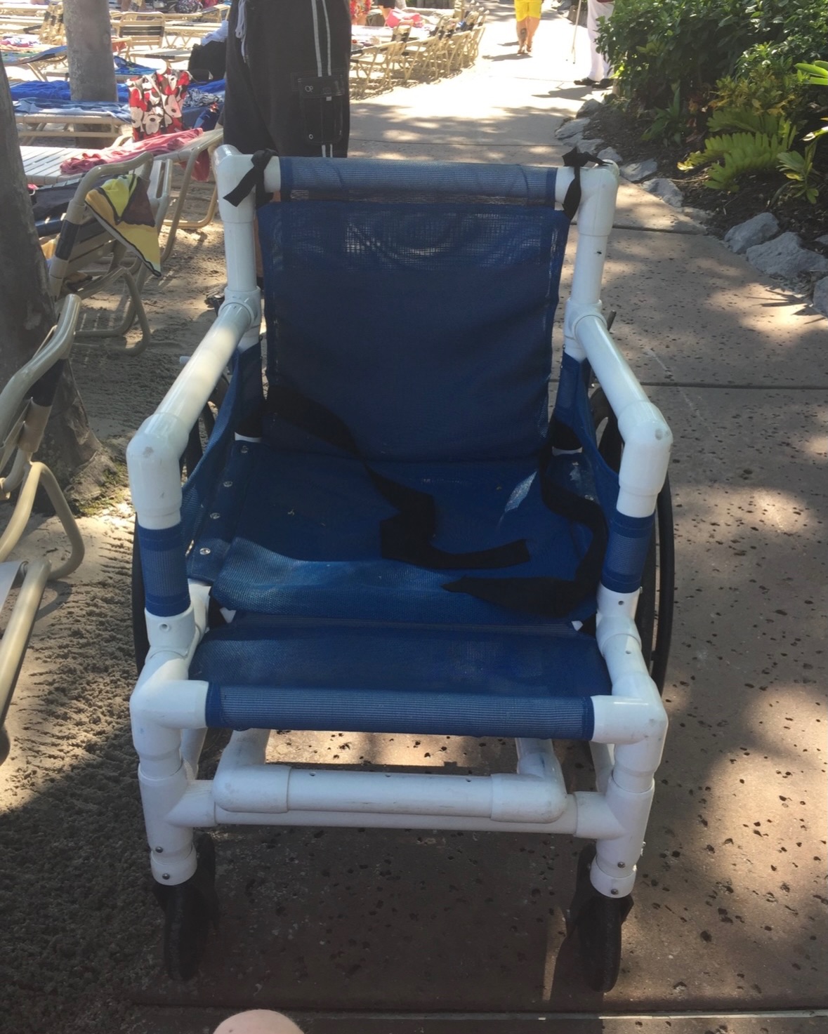 A beach wheelchair with a white PVC pipe frame, blue mesh seat and backrest, black straps and black wheels, parked on a shaded concrete path beside sand and rows of lounge chairs