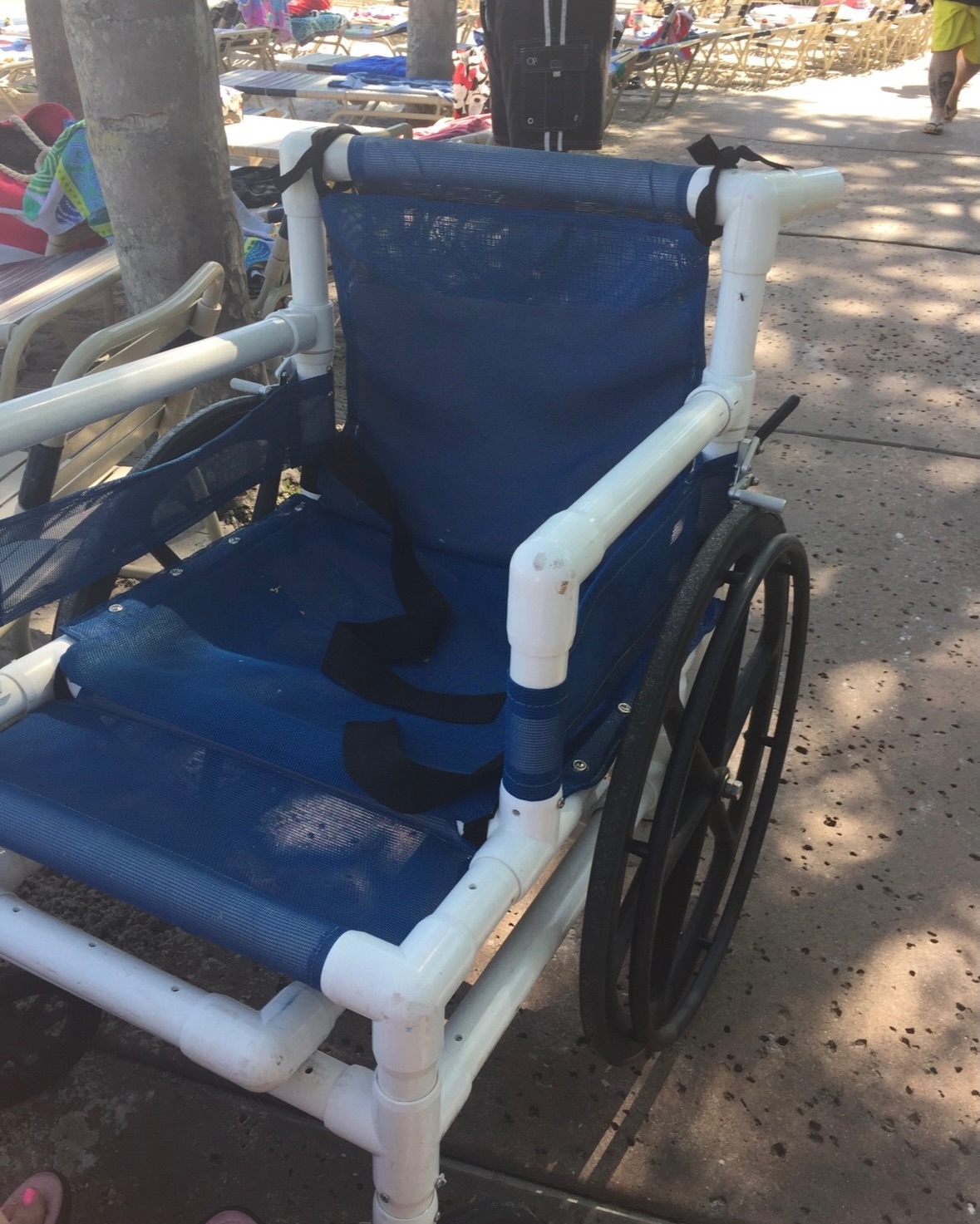 The same blue mesh and white PVC beach wheelchair seen from an angle, showing its large black rear wheel, on a shaded path with lounge chairs and tree trunks behind it