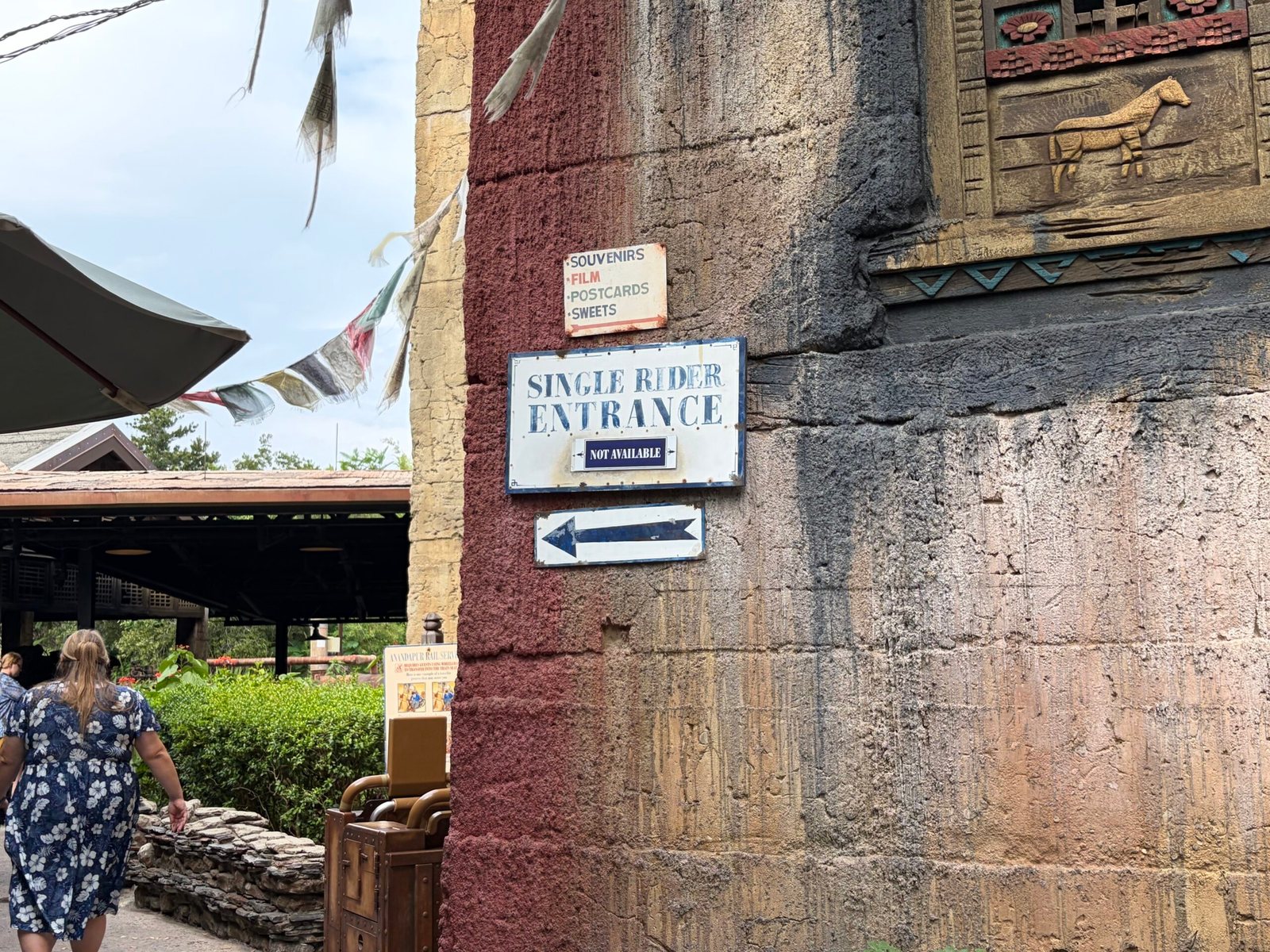 A weathered Single Rider Entrance sign with a small Not Available placard and a left-pointing arrow beneath it, mounted on a red and gray rock wall below a Souvenirs, Film, Postcards, Sweets sign, with prayer flags and a guest walking past at left