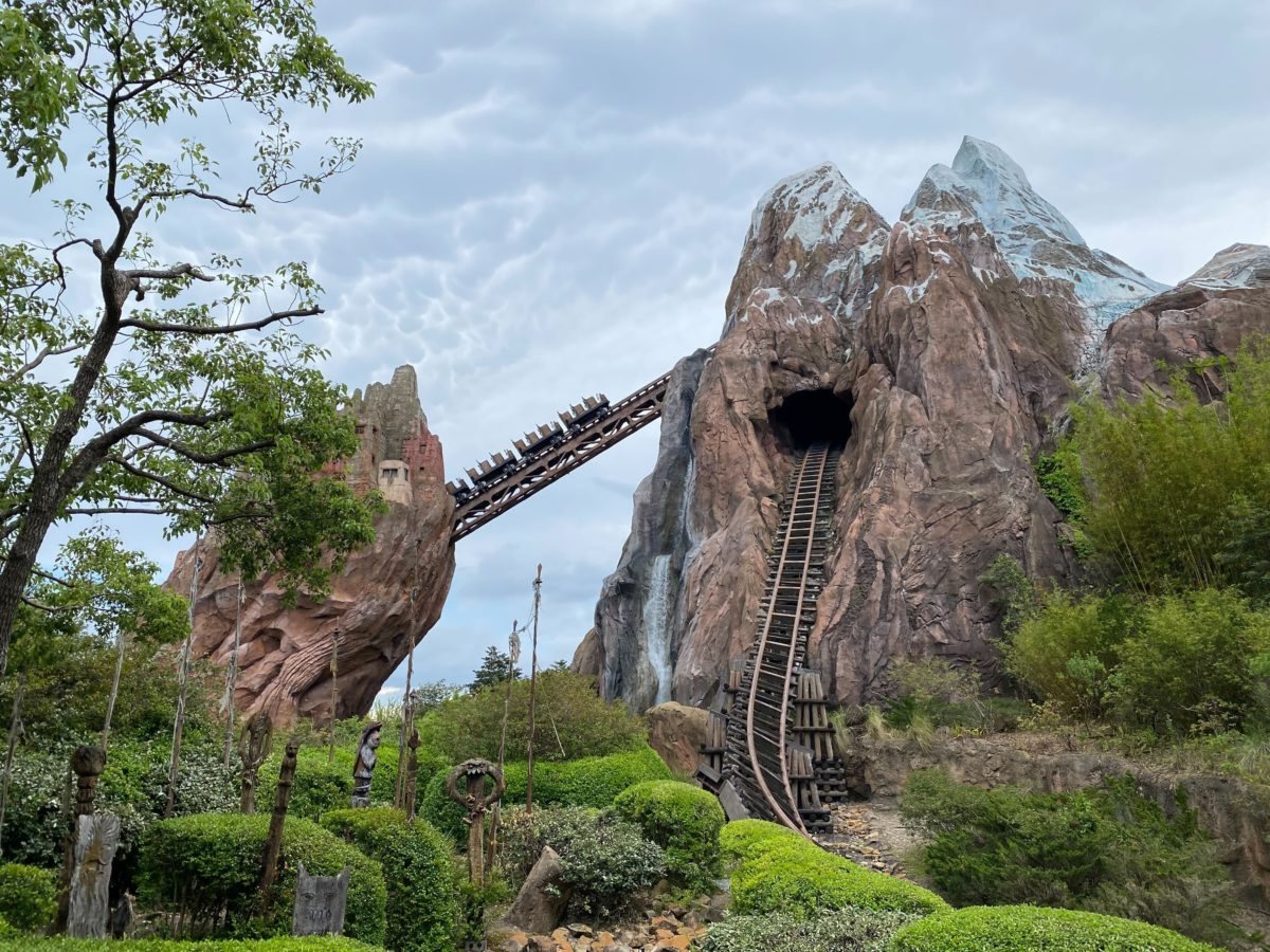 Expedition Everest's snowcapped rockwork mountain at Disney's Animal Kingdom under a cloudy sky, with a train on the elevated track and the ride's track disappearing into a tunnel in the mountain