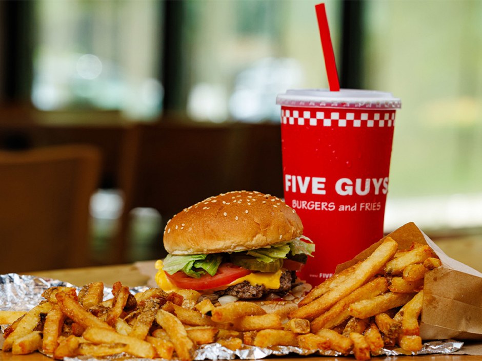 A cheeseburger with lettuce, tomato and pickles beside a pile of fries on foil and a red Five Guys Burgers and Fries cup with a straw