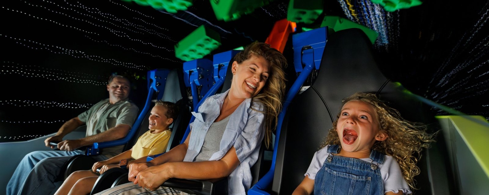A family laughing and screaming on a dark indoor roller coaster with blue restraints, as giant green and orange LEGO bricks hang overhead