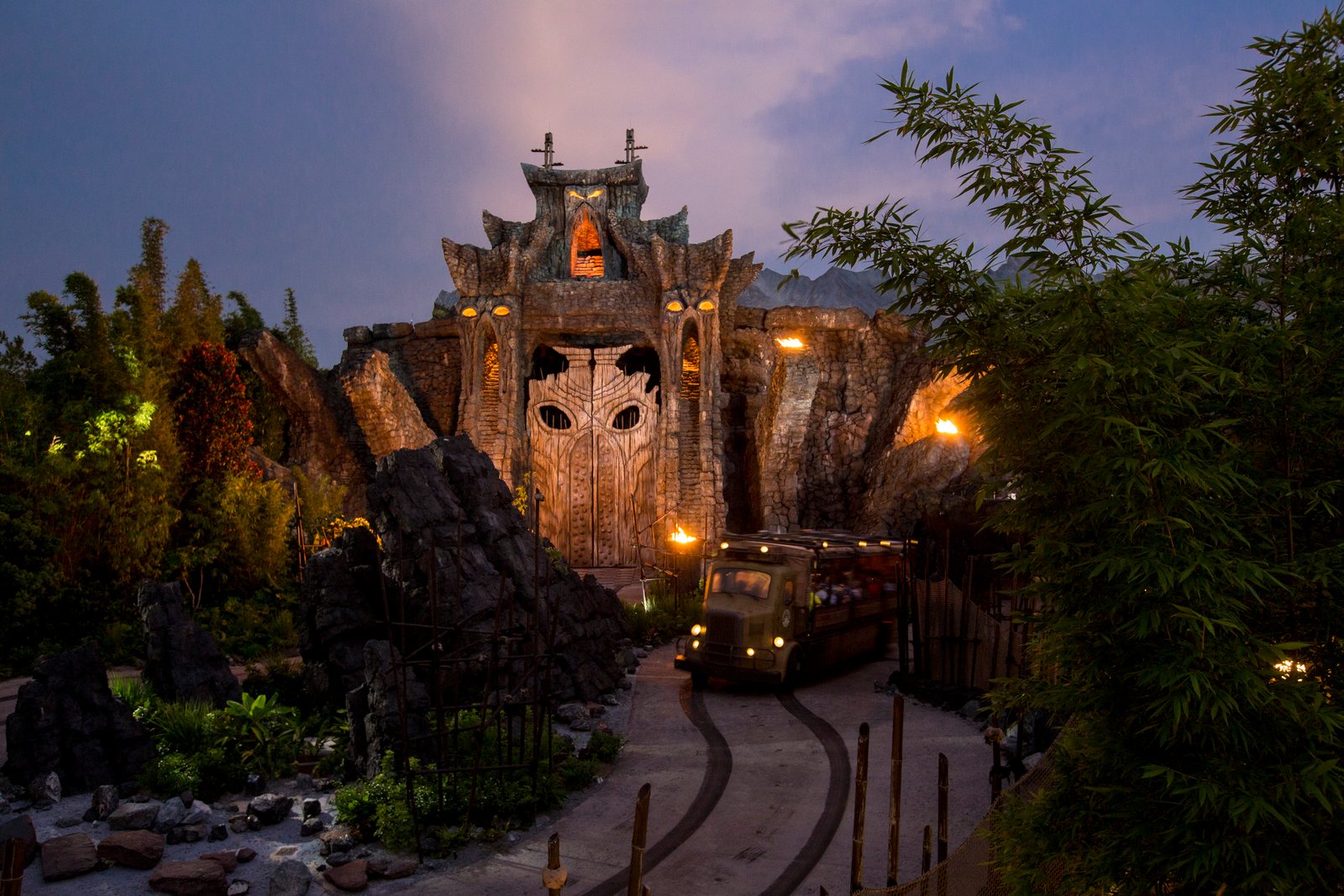 The carved wooden temple gate of Skull Island: Reign of Kong at dusk, lit by torches, with a green expedition truck carrying riders along the outdoor path