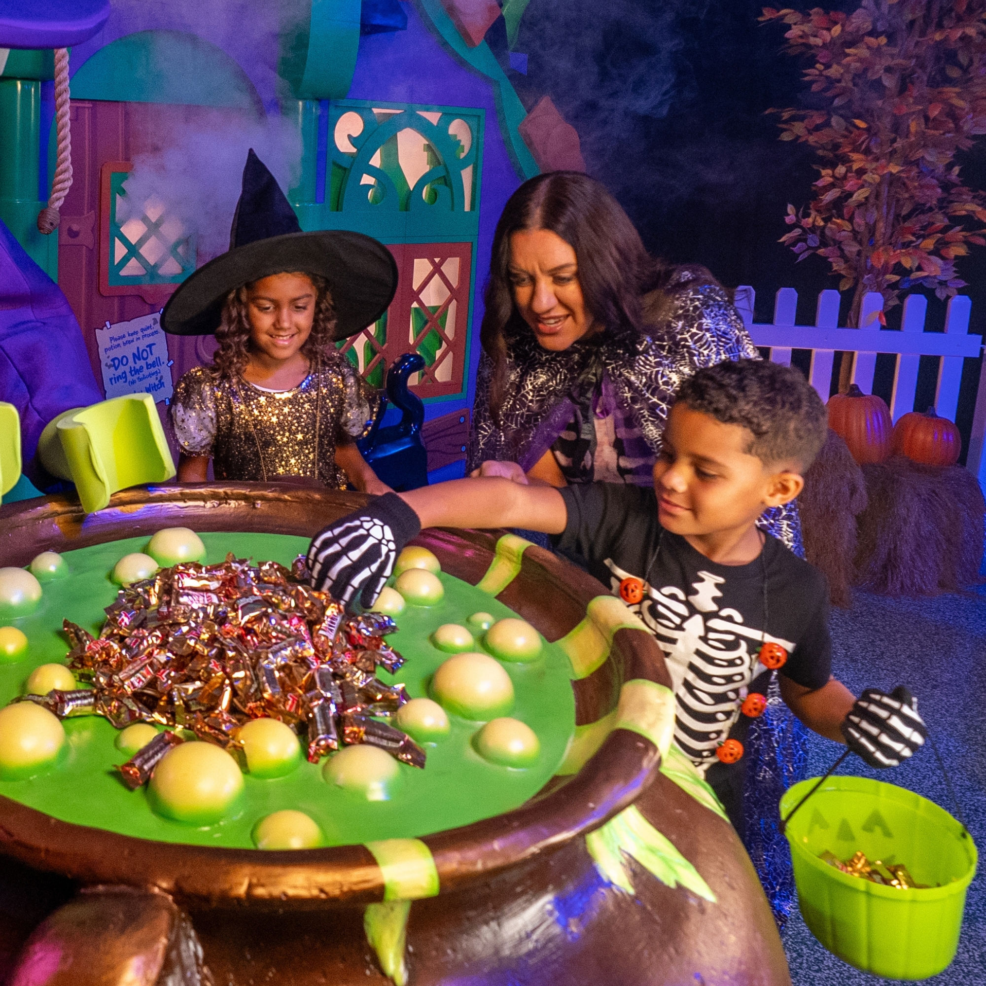 A boy in a skeleton costume reaching into a large cauldron of green potion and wrapped candy while a girl in a witch hat and a costumed witch character look on, outside a purple and teal Lego cottage facade with fog, a picket fence and pumpkins behind them