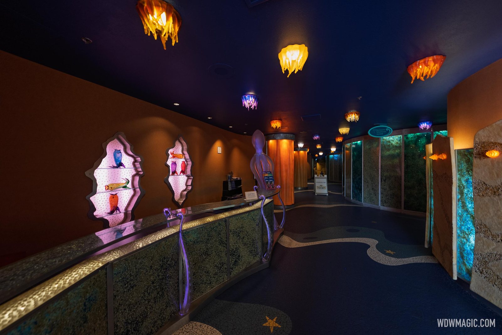 The dimly lit Coral Reef Restaurant check-in counter, with glass jellyfish light fixtures on the ceiling, glass fish sculptures in wall niches and a wavy mosaic floor
