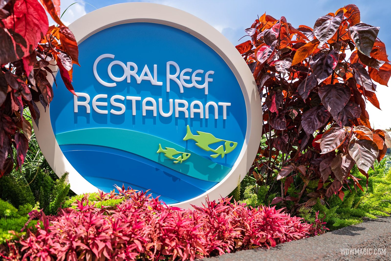 The round blue Coral Reef Restaurant sign with two green fish, surrounded by red and pink plants under a blue sky
