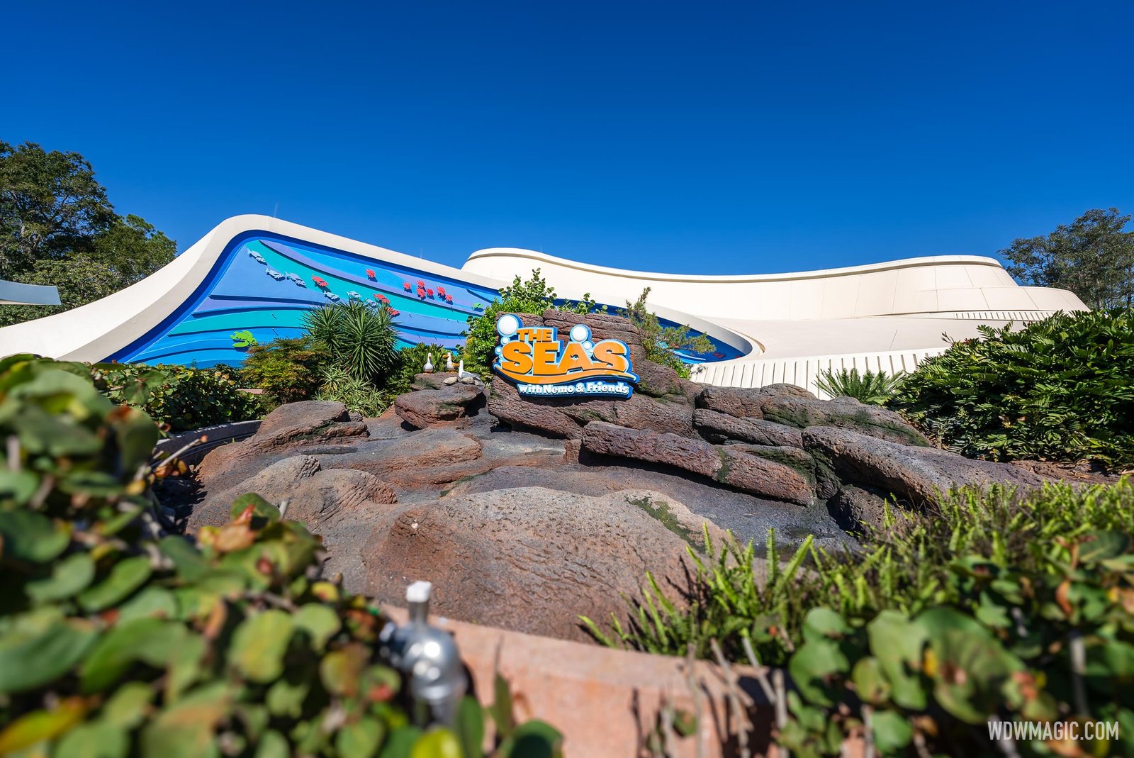 The curving white and blue exterior of The Seas with Nemo & Friends pavilion at EPCOT behind its rockwork sign, under a clear blue sky
