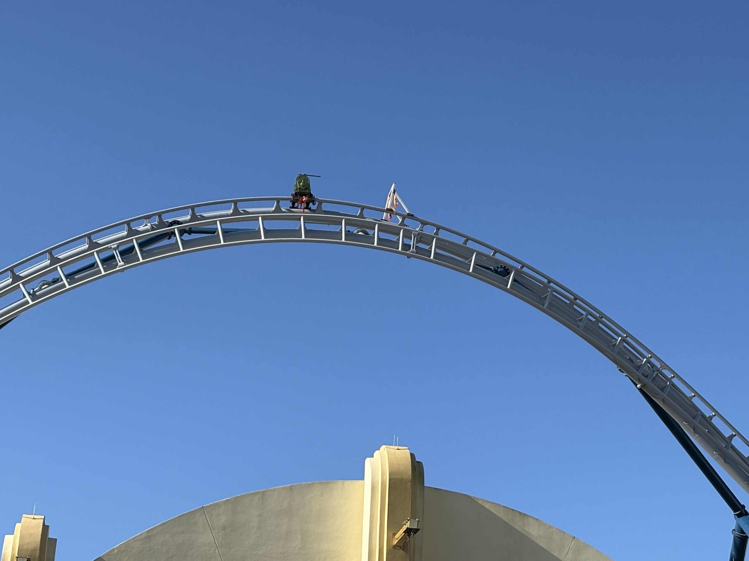 A green spool apparatus mounted on the crest of a gray coaster track arch against a clear blue sky, secured with an orange mounting assembly, with a warning flag beside it and a beige stage structure below