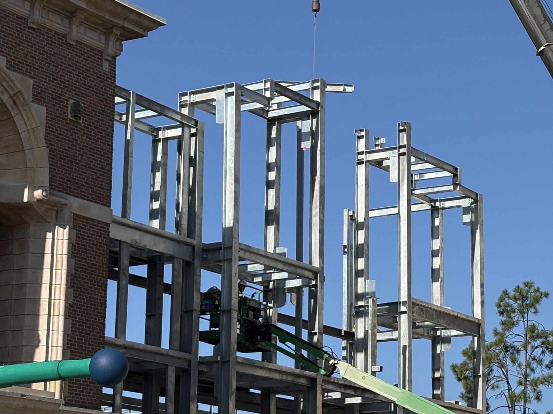 Tall galvanized steel framework of rectangular bays with no walls or roof, rising beside an existing brick facade with stone detailing and a large arched opening, a green boom lift working inside the frame