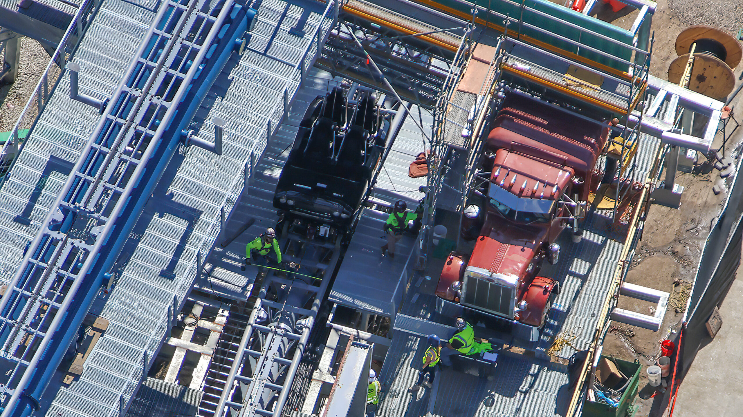 Aerial view looking straight down at a black ride vehicle sitting on coaster track beside a burgundy truck cab prop, with workers in high visibility vests on metal grating around them