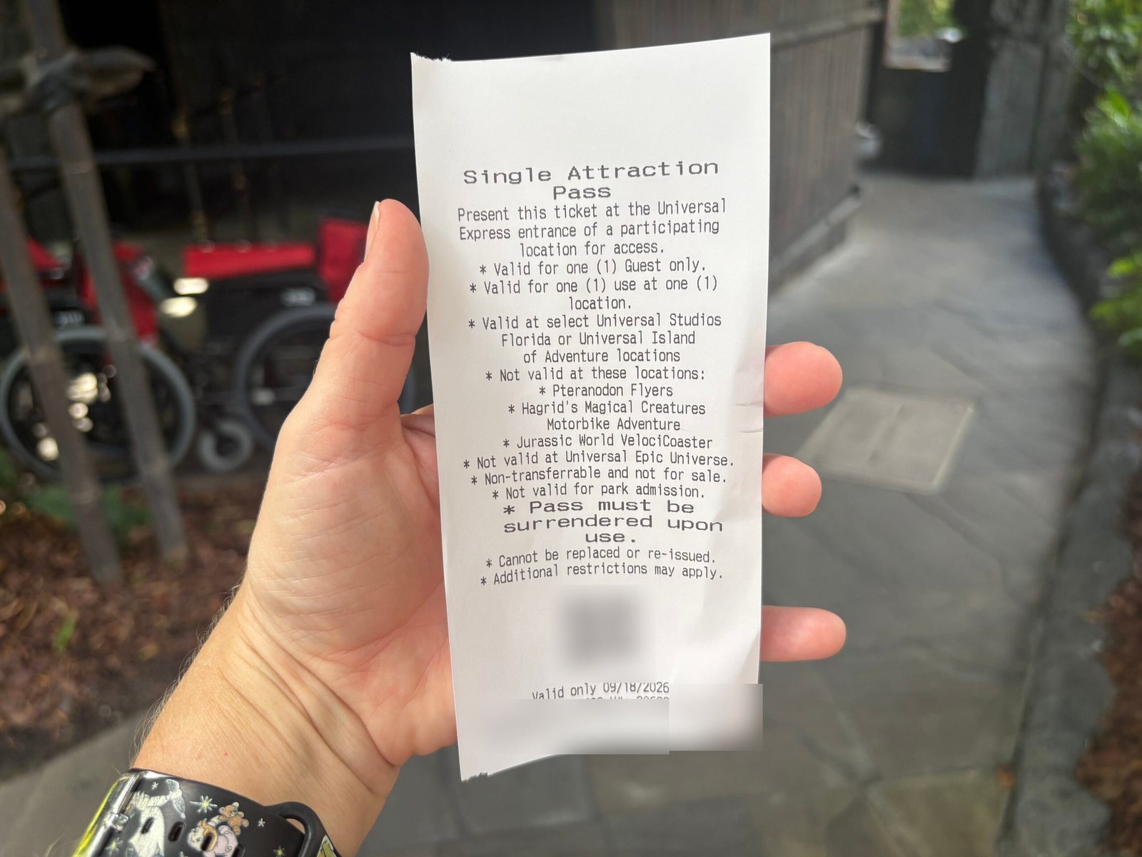 A hand holds a printed receipt-style Single Attraction Pass listing its rules, including that it is not valid at Pteranodon Flyers, Hagrid's Magical Creatures Motorbike Adventure, Jurassic World VelociCoaster or Universal Epic Universe