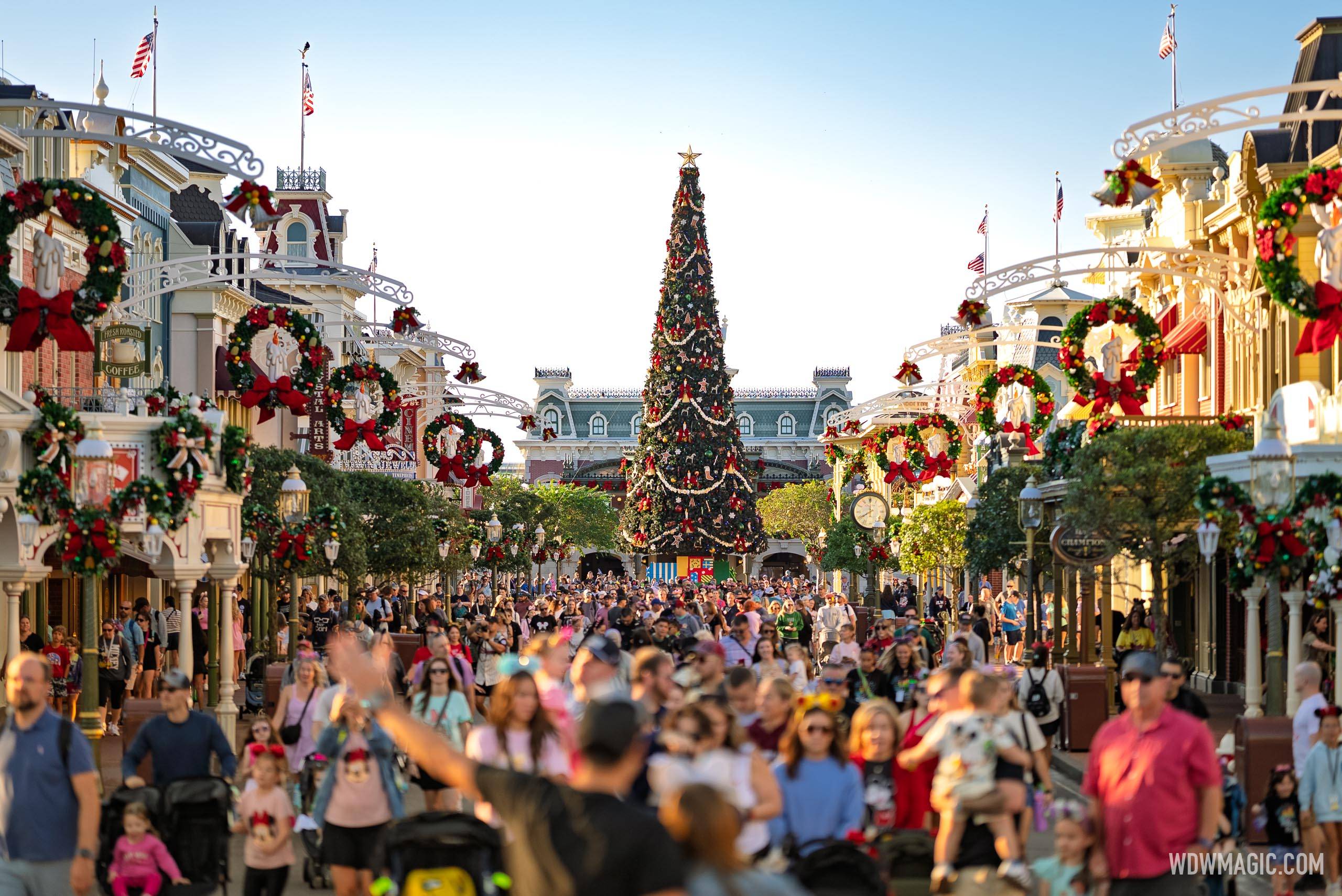 Main Street, U.S.A. packed with guests in daylight, looking toward the train station with the tall Magic Kingdom Christmas tree decorated with garlands and gingerbread ornaments in the center