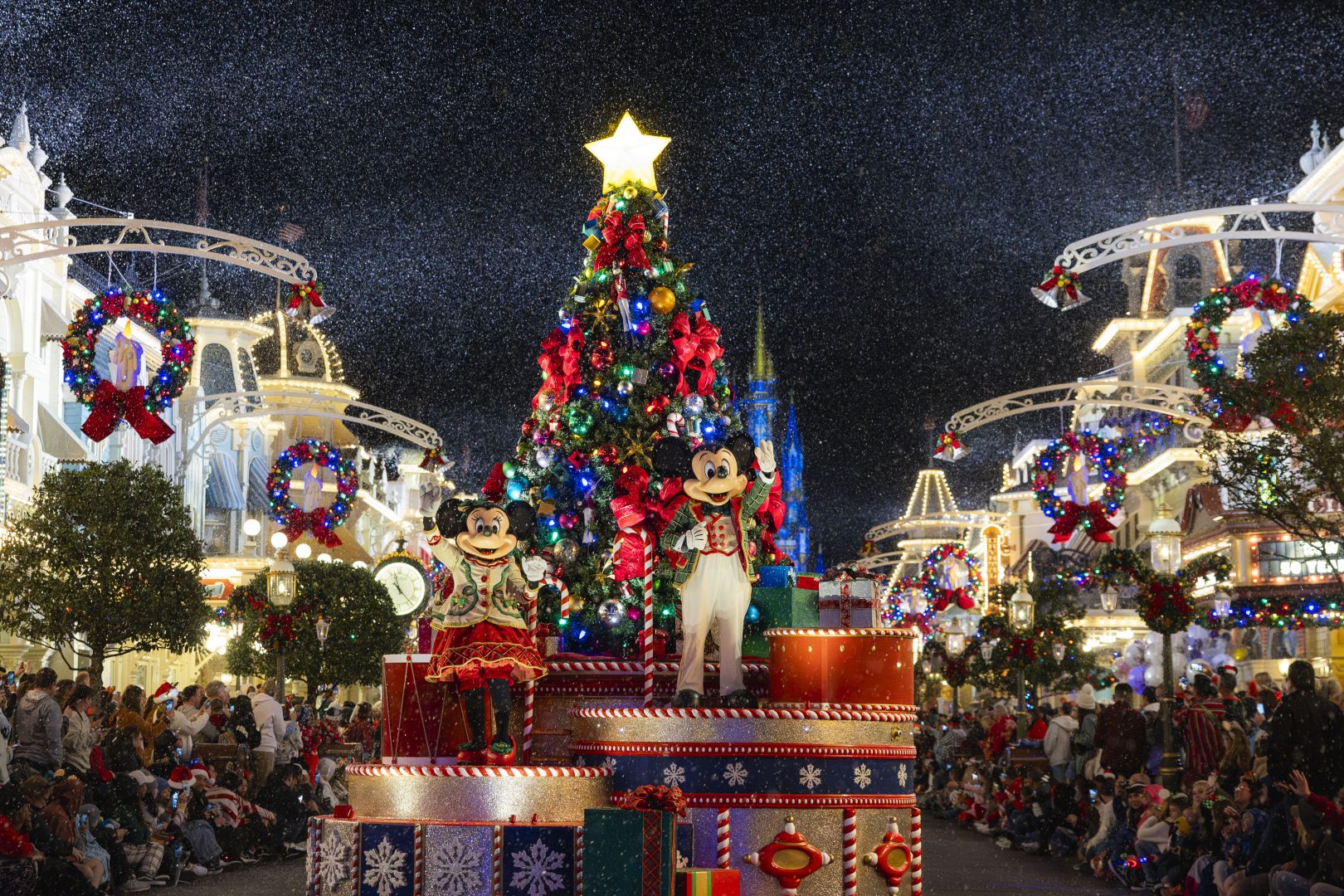 Mickey and Minnie Mouse in holiday outfits waving from a glittered parade float built of oversized wrapped presents on Main Street at night, with a lit Christmas tree behind them and Cinderella Castle glowing blue, artificial snow falling