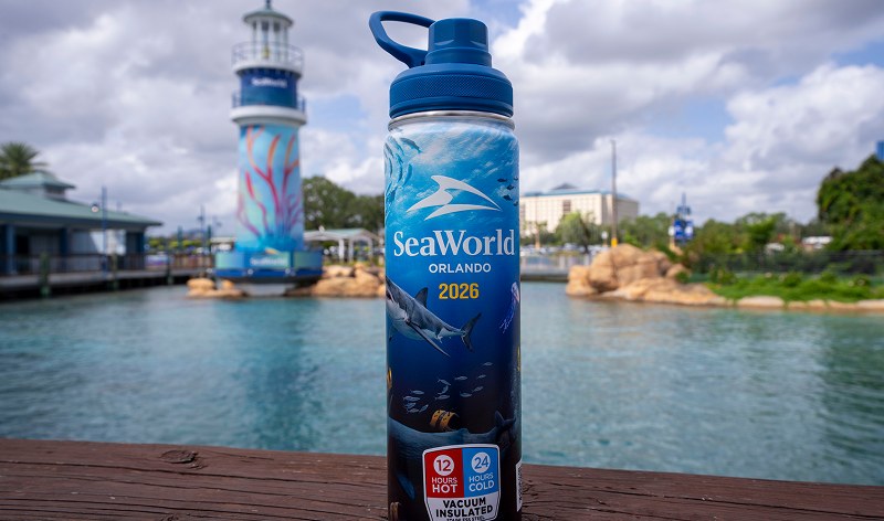 A tall navy vacuum insulated bottle standing on a weathered wood rail in front of a blurred lagoon, wrapped in underwater art with a shark and reading SeaWorld Orlando 2026, with a badge reading 12 hours hot 24 hours cold vacuum insulated