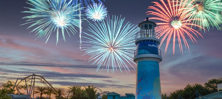 Dusk over SeaWorld with teal, white, red and green firework bursts filling a pink and blue sky, the blue SeaWorld lighthouse at center right and a coaster track and palms at left