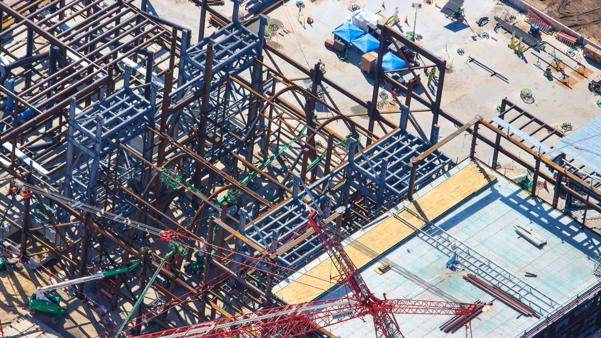 Close aerial of the steel framing showing curved steel members among the straight grid, green lifts working at height, and a run of corrugated decking with plywood at lower right