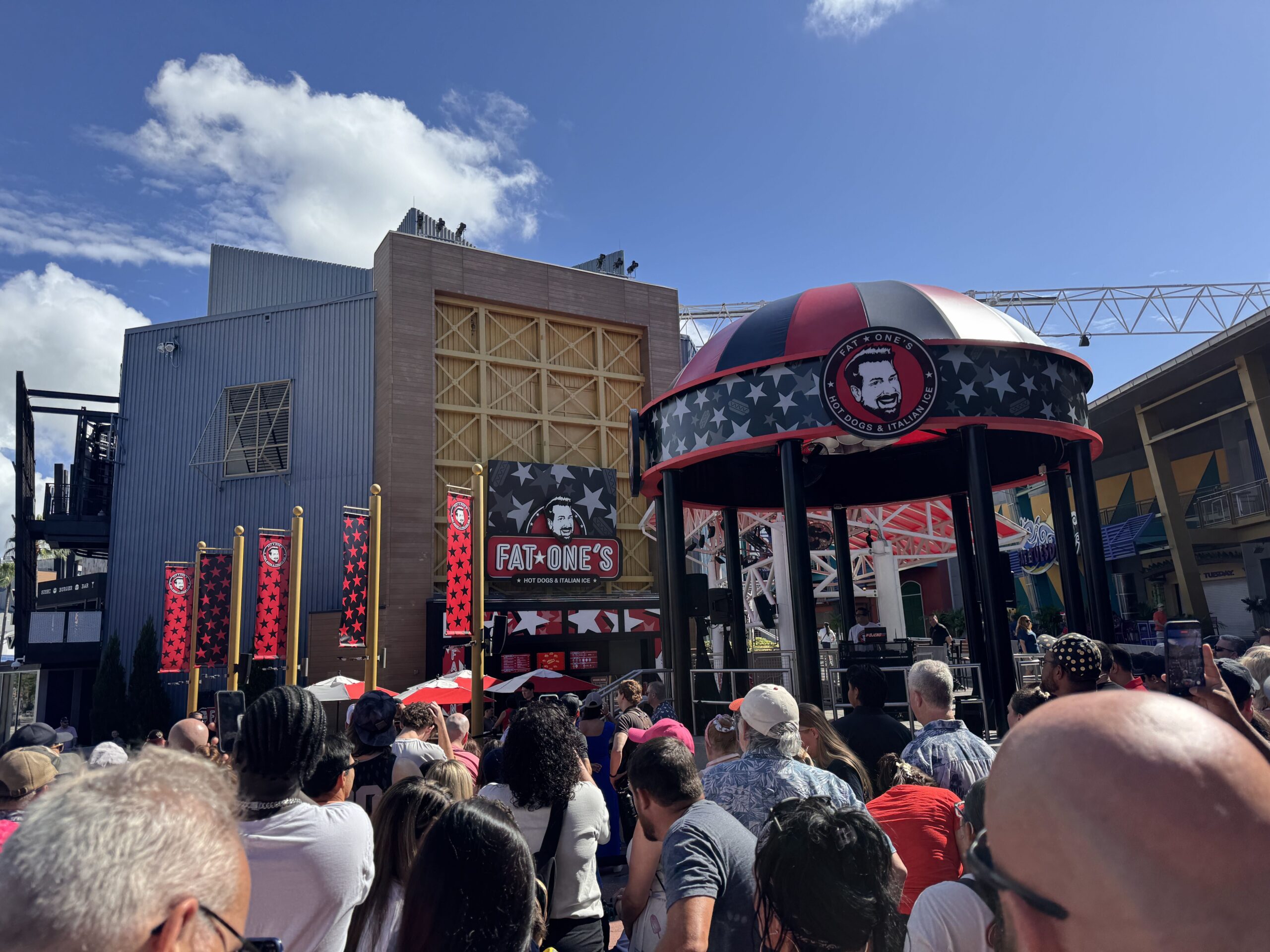 The back of a dense crowd of guests filming on phones at CityWalk, facing a circular bandstand topped with a red, black and white striped dome carrying the round Fat One's logo, with the restaurant facade and red star banners at left