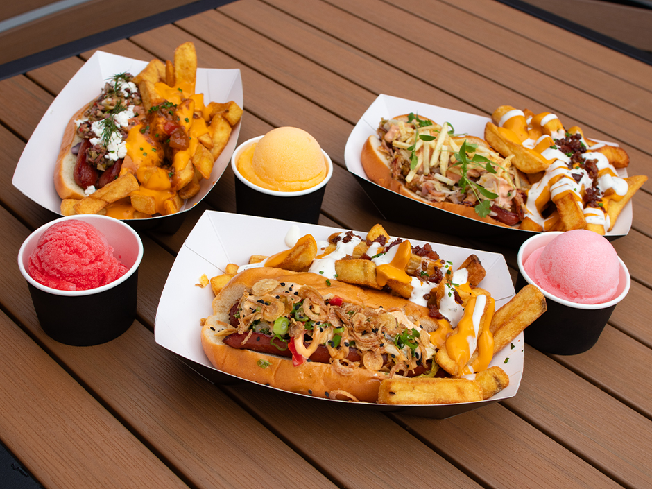 Three loaded hot dogs in black and white paper boats, each sharing its boat with a pile of loaded fries, arranged on a slatted brown patio table with three black cups of Italian ice in red, orange and pink between them