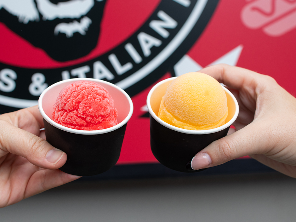 Two hands holding up two small black paper cups of Italian ice, one scoop bright red and one orange, in front of a red Fat One's logo sign that is slightly out of focus