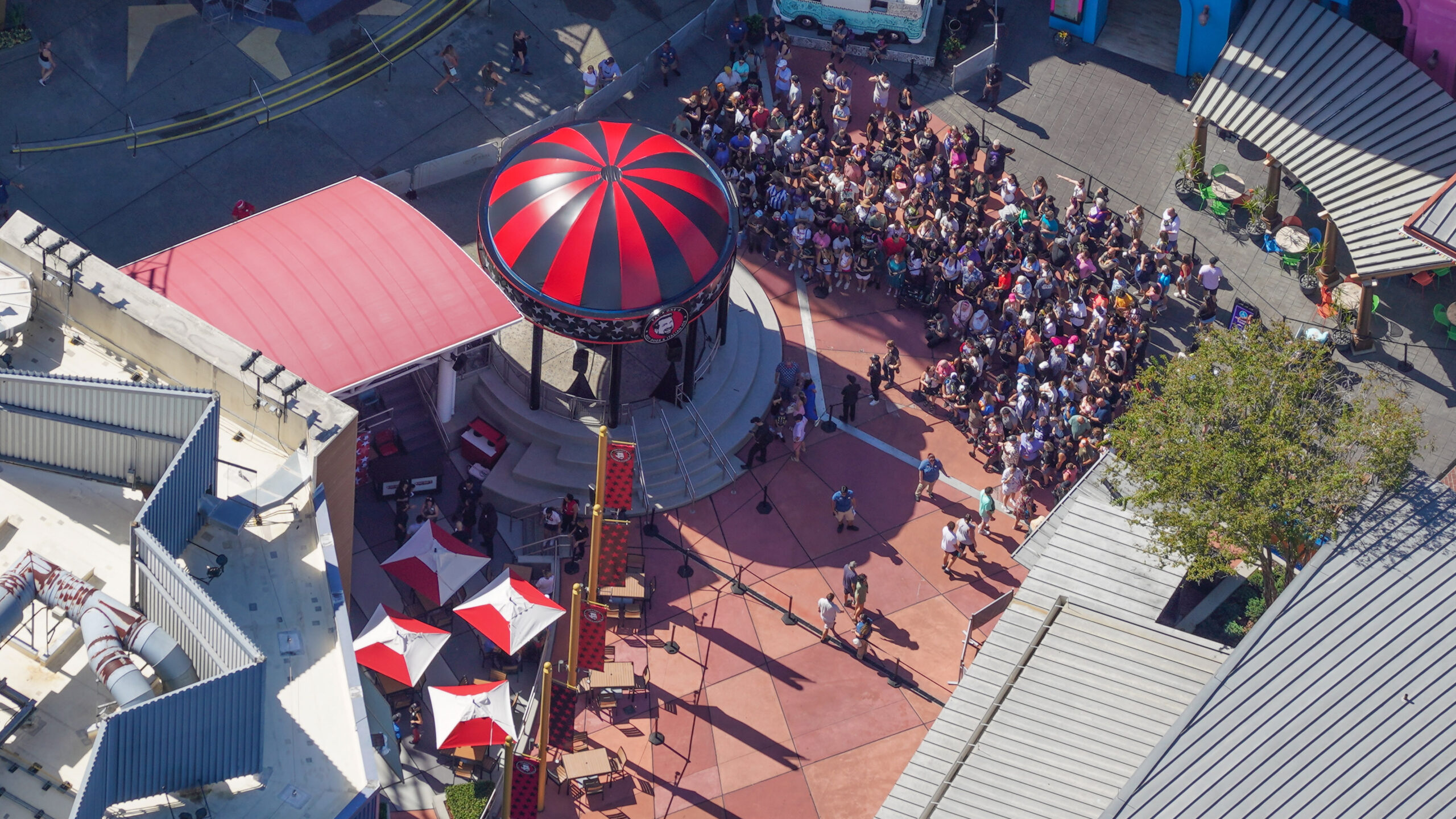 High angle aerial view of the CityWalk plaza showing the red and black striped bandstand dome in the center, several hundred guests packed behind stanchion lines to its right, the restaurant's red roof at left and red and white umbrella tables over its patio at lower left