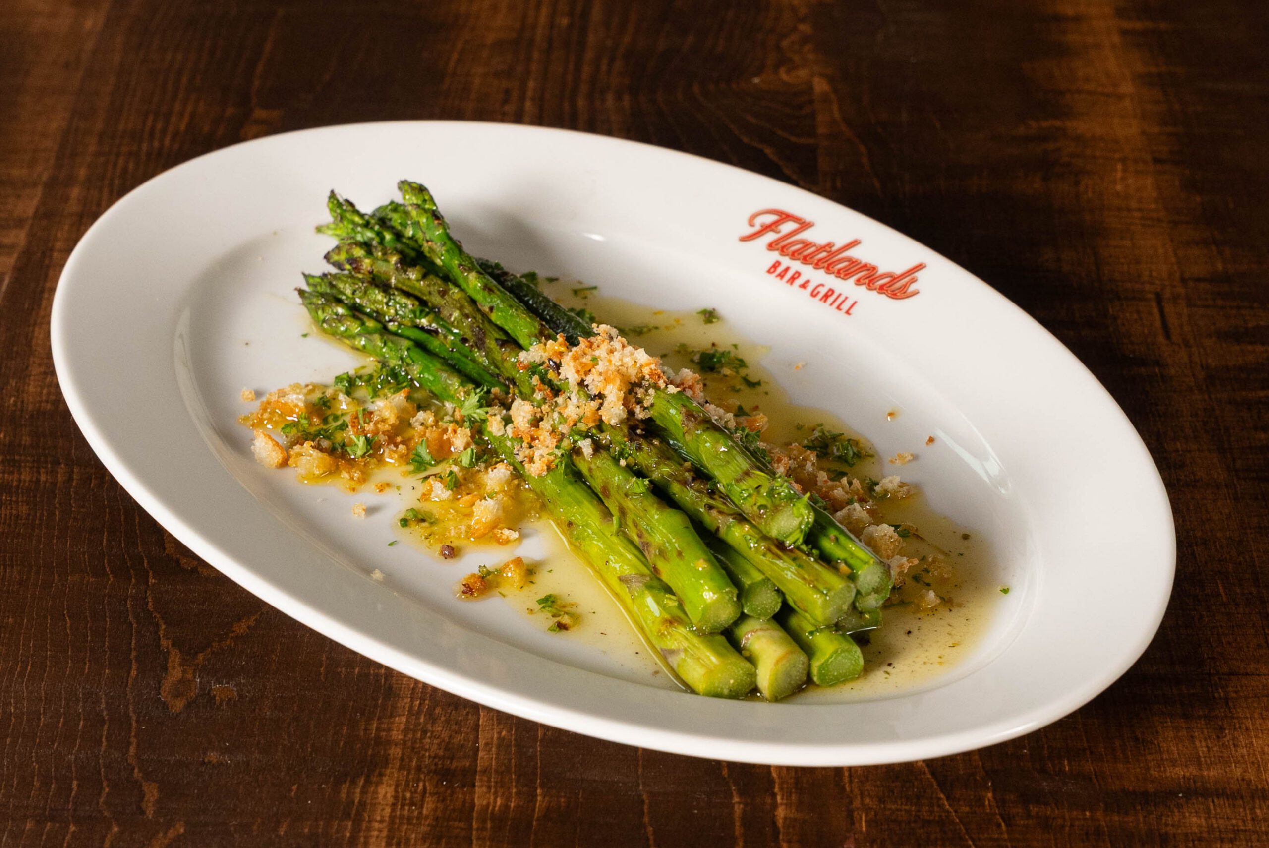 Grilled asparagus spears on a white oval plate set on dark wood, topped with toasted breadcrumbs, chopped parsley and a glossy lemon and olive oil dressing, with the red Flatlands Bar & Grill logo, script over capitals, printed on the upper right rim of the plate