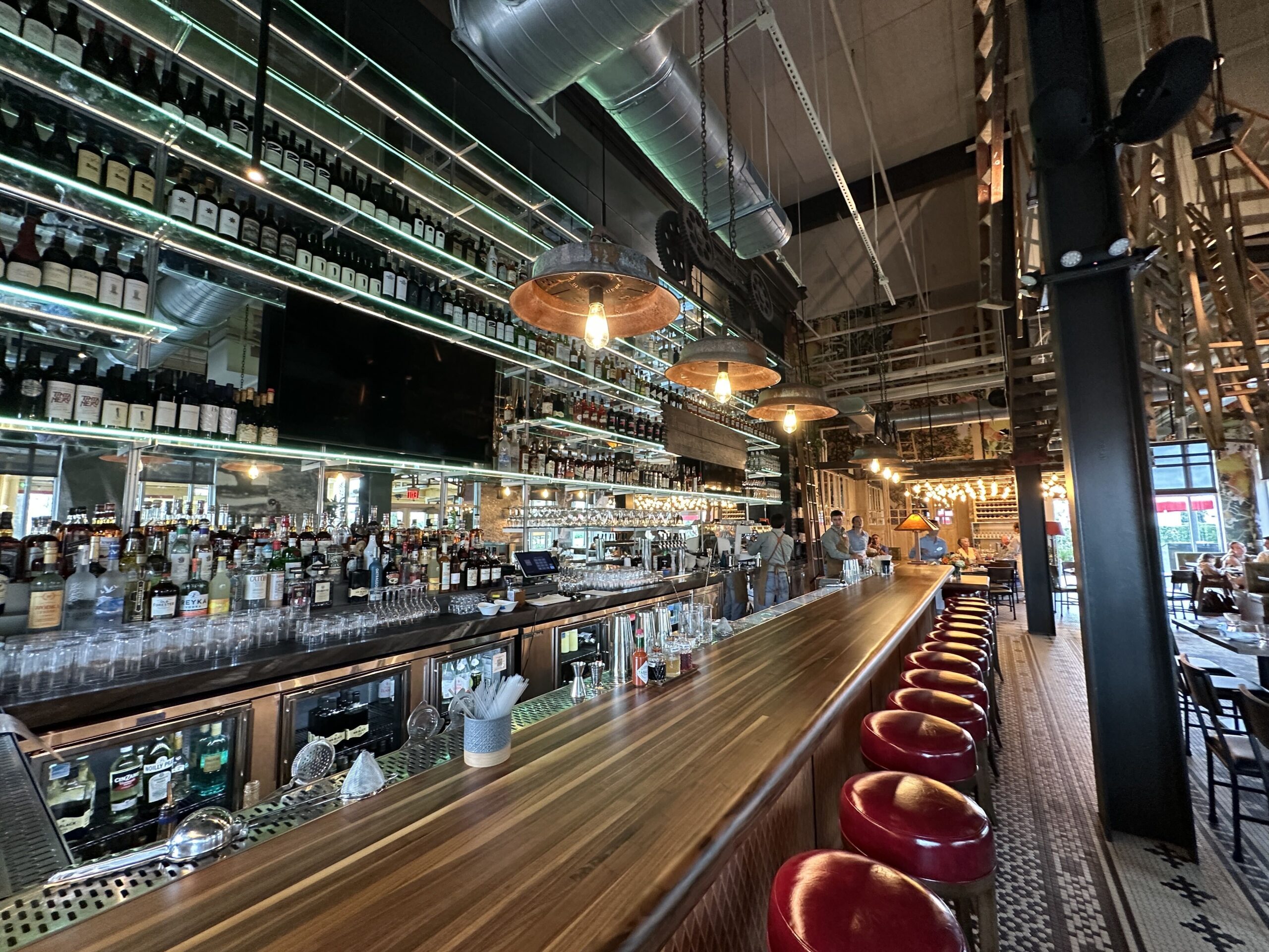 A long polished hardwood bar top running into the distance with oxblood red leather stools along it, backlit glass shelving stacked several tiers high with liquor and wine bottles behind it, rusted metal pendant lamps with exposed bulbs, silver ductwork and wooden ladders hung at angles near the ceiling