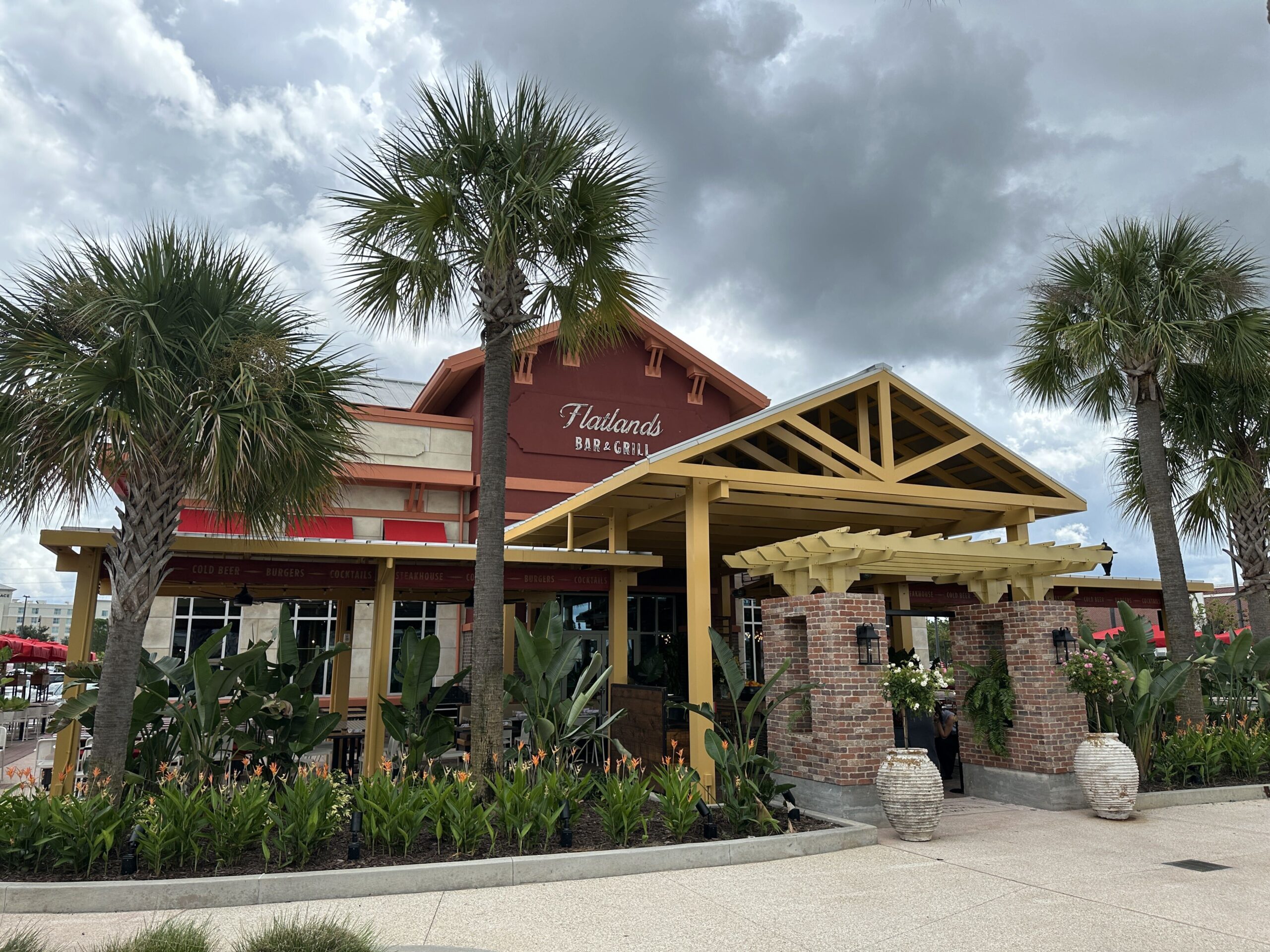 A deep red gabled restaurant building reading Flatlands in white script above BAR & GRILL in white capitals, with a mustard yellow timber porte cochere and pergola in front, red brick piers with lanterns, a red awning band reading COLD BEER BURGERS COCKTAILS STEAKHOUSE, and sabal palms and broad-leaved tropical plantings with orange flowers around it under an overcast sky
