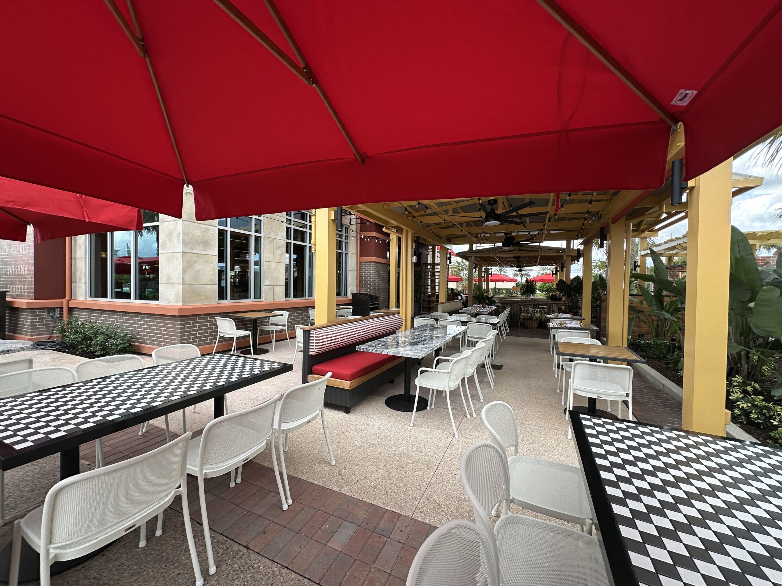 A covered outdoor patio under a large red umbrella, with black and white checkerboard topped tables, white mesh chairs, a red cushioned banquette beside a grey marble communal table, yellow posts, ceiling fans and string lights overhead and tropical plantings along the right side